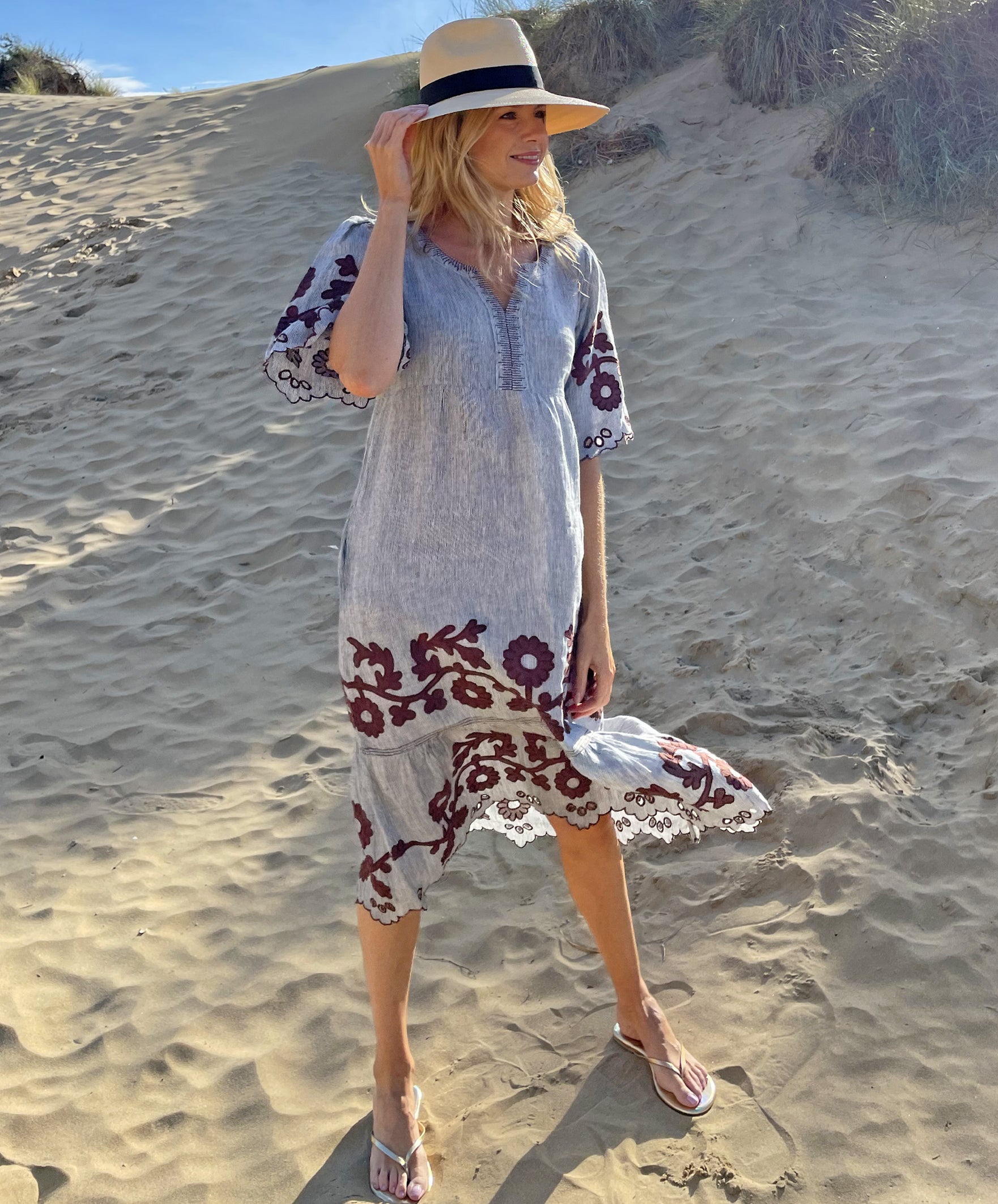 A model stood on the beach holding onto her hat and wearing a Rose and Rose striped linen Agrigento dress.