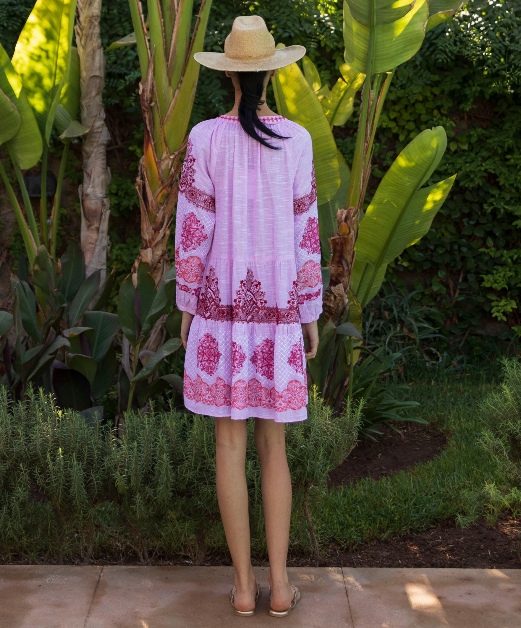Back view of a woman wearing a pink embroidered Alder dress in a tropical garden setting.