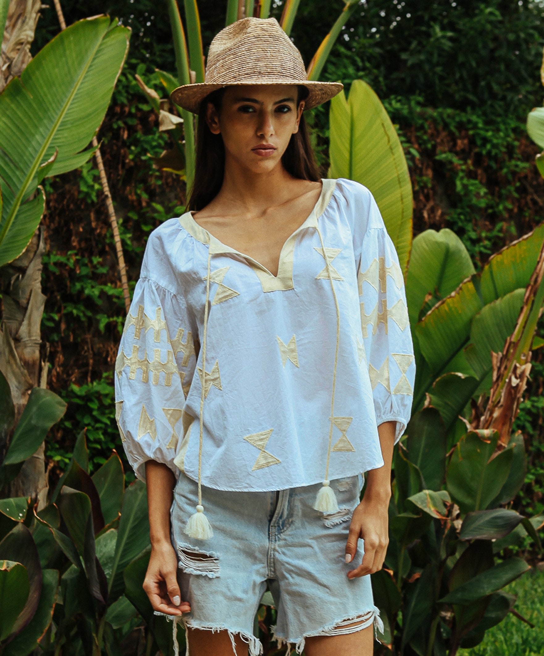 A model stood in a tropical garden wearing a white Rose and Rose Aldgate cotton top with gold lurex appliqué decoration, a sun hat, and denim shorts.