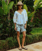 A model stood in a tropical garden wearing a white Rose and Rose Aldgate cotton top with gold lurex appliqué decoration, a sun hat, and denim shorts.