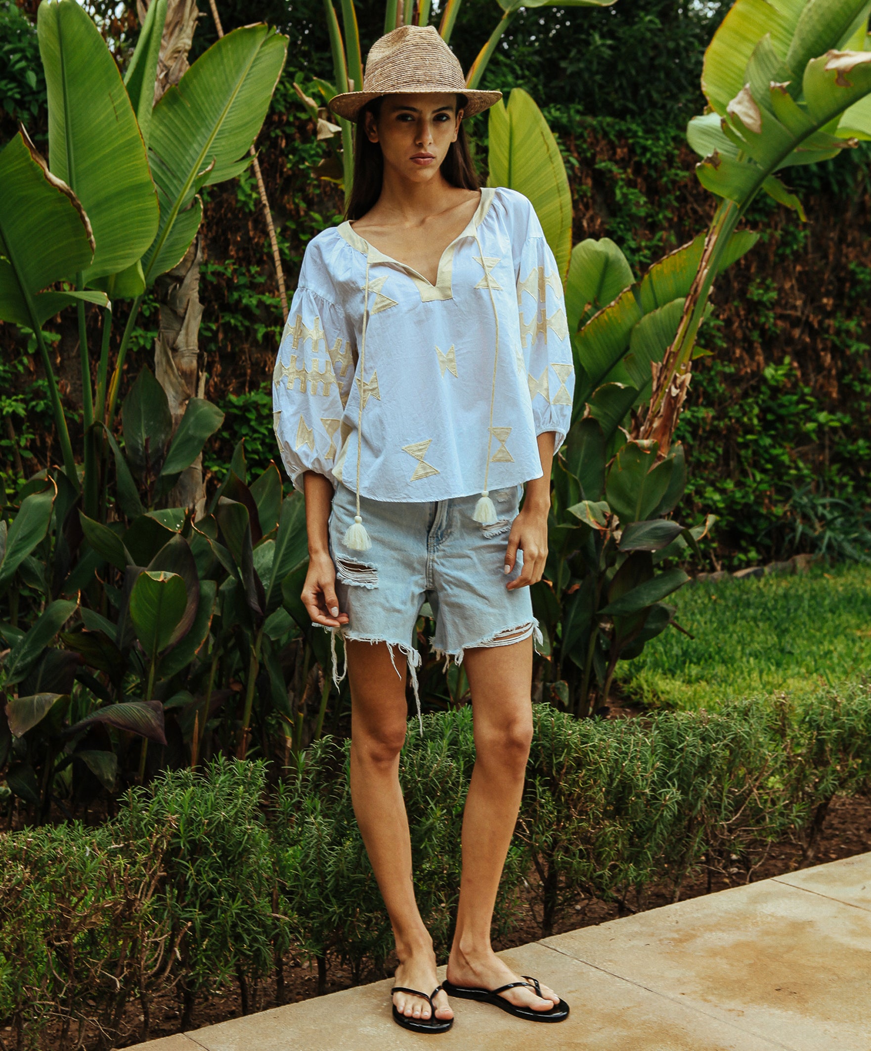 A model stood in a tropical garden wearing a white Rose and Rose Aldgate cotton top with gold lurex appliqué decoration, a sun hat, and denim shorts.