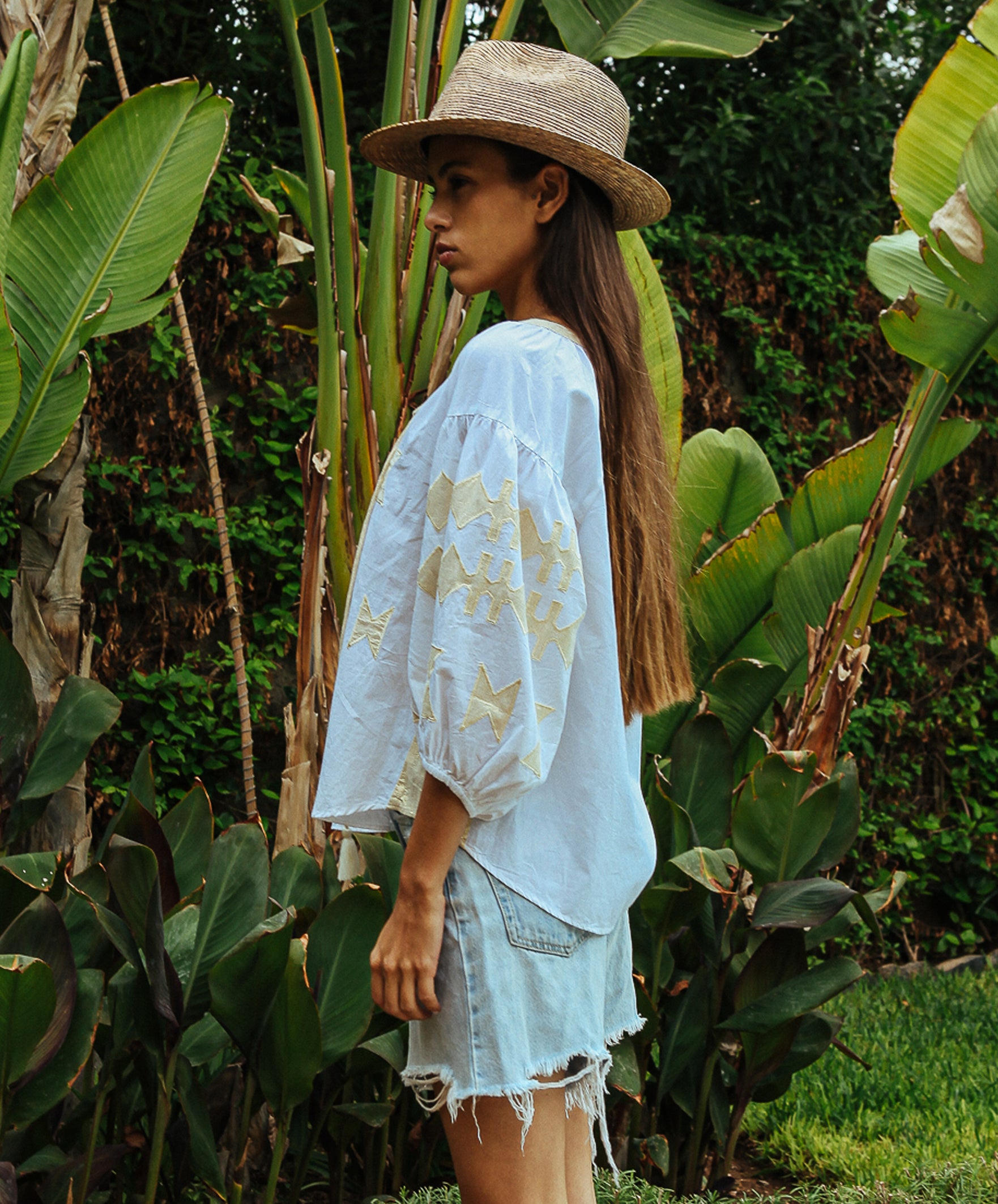 Side view of a model stood in a tropical garden wearing a white Rose and Rose Aldgate cotton top with gold lurex appliqué decoration, a sun hat, and denim shorts.