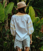Back view of a model stood in a tropical garden wearing a white Rose and Rose Aldgate cotton top with gold lurex appliqué decoration, a sun hat, and denim shorts.