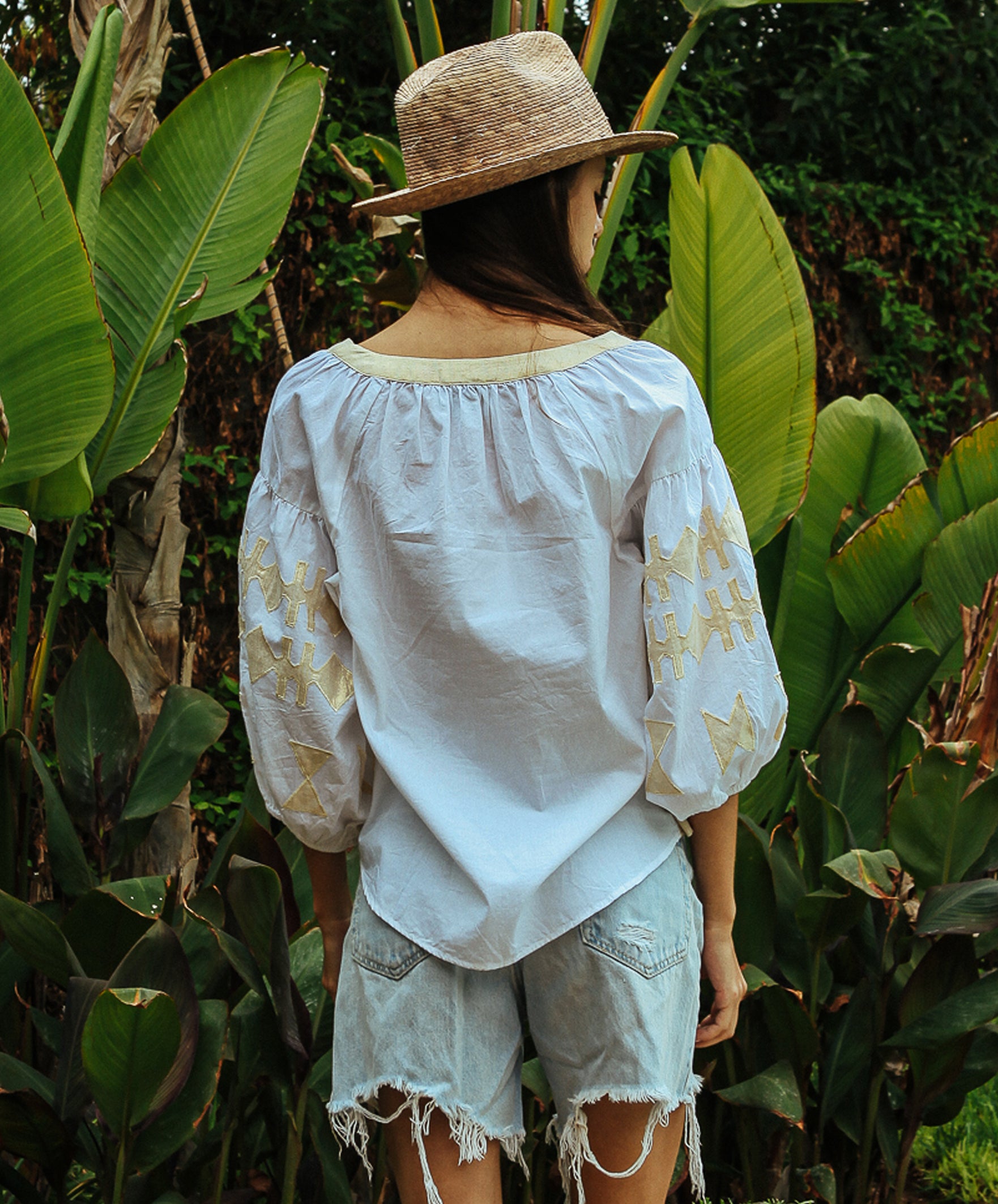 Back view of a model stood in a tropical garden wearing a white Rose and Rose Aldgate cotton top with gold lurex appliqué decoration, a sun hat, and denim shorts.