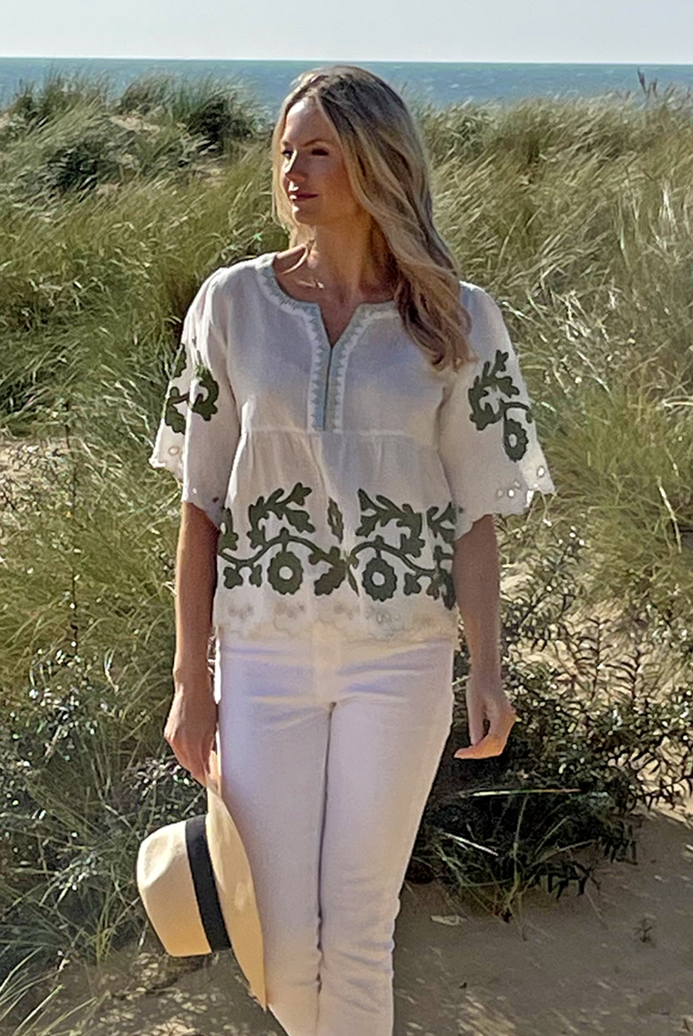 A model on a beach wearing the Rose and Rose Alessio top in white linen with olive applique and holding an Anthony Peto Panama hat. 