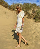 A side view of a model on a beach wearing the Rose and Rose Amalfi dress in gold lurex with white applique.
