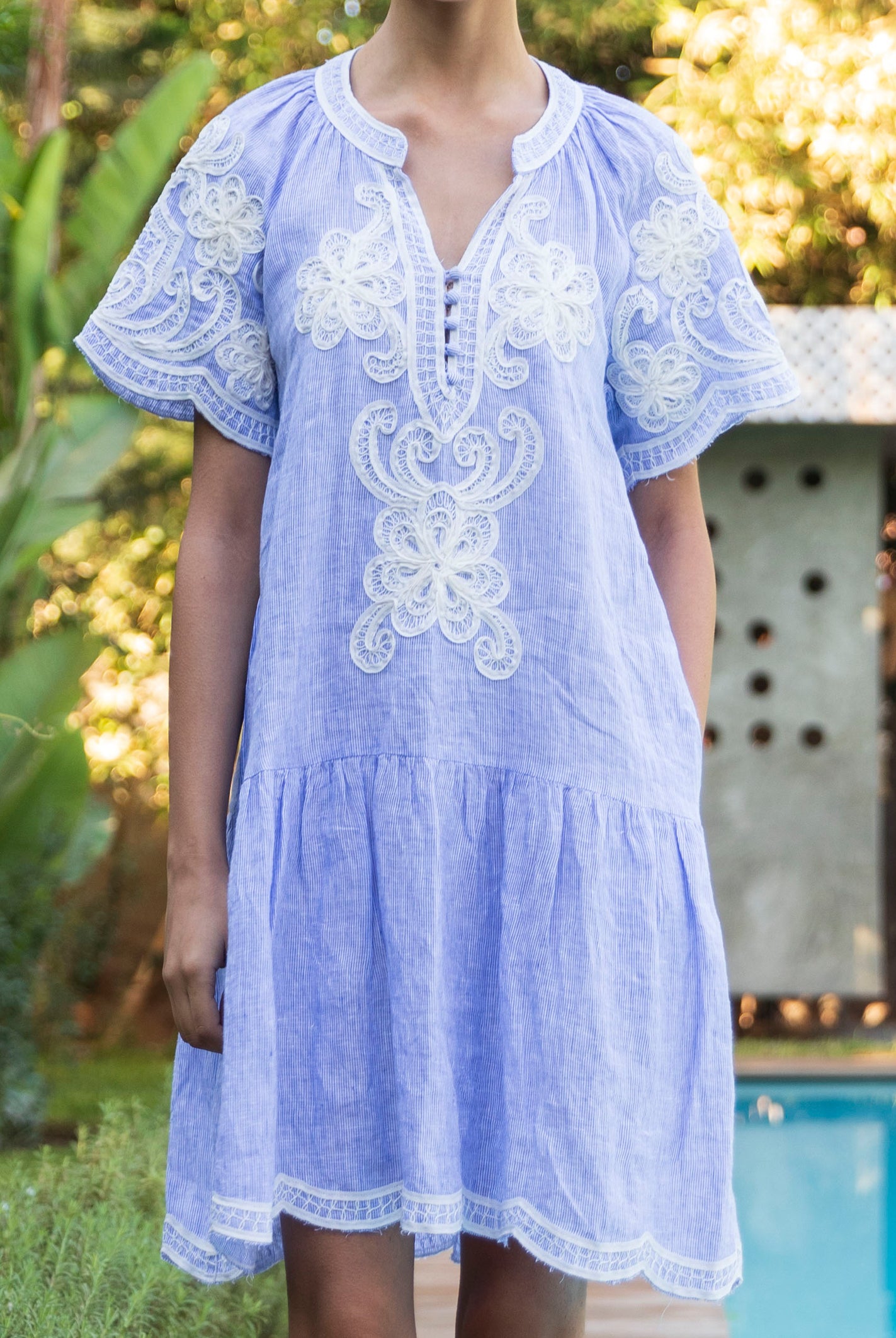 Striped blue Amaranta dress with white floral tapework decoration worn by a pool.