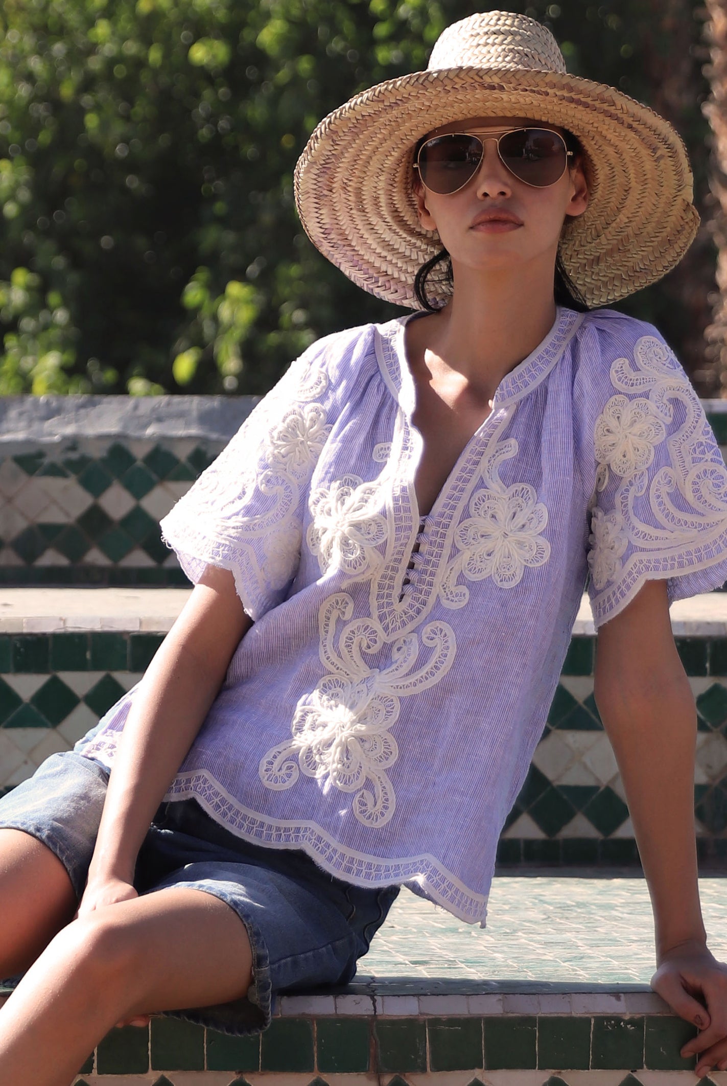 Woman sat on steps wearing a blue striped embroidered Azalea top and straw hat