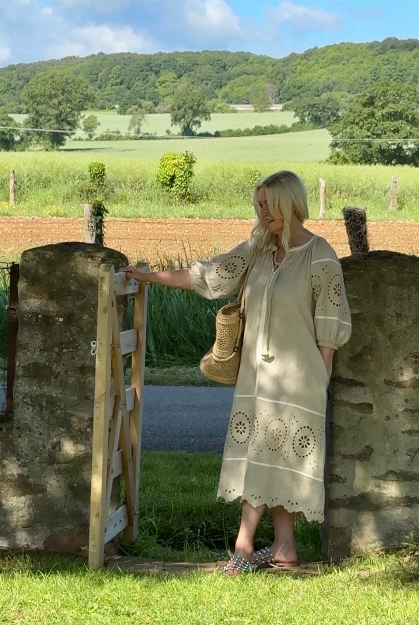 Influencer @shutters_and_stone stood wearing the Magnolia natural embroidered dress standing next to a stone gate with a scenic background of fields and trees.