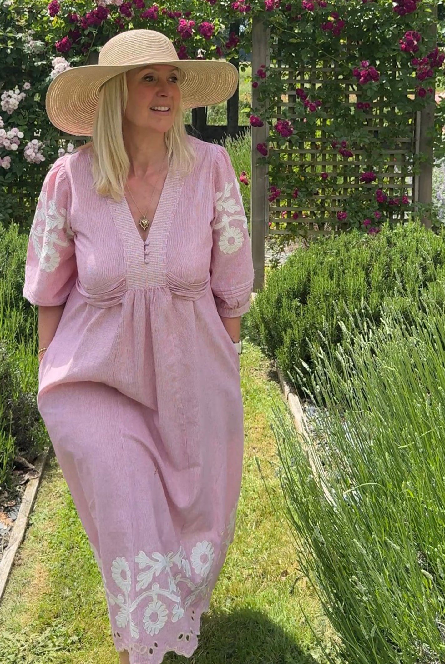 Influencer @shutters_and_stone wearing the Begonia pink dress and hat standing in a garden with lavender and flowers.