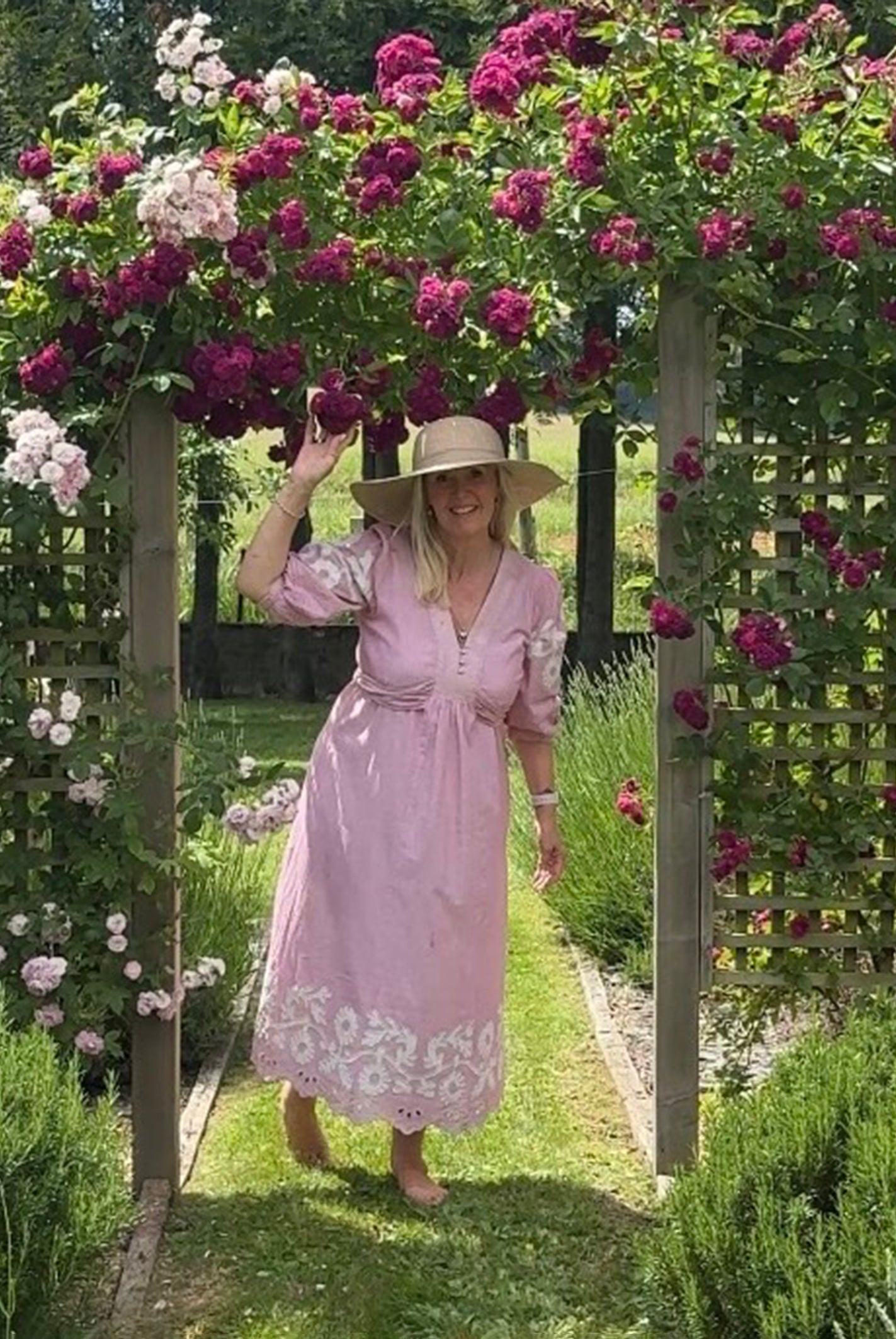 Influencer @shutters_and_stone wearing the Begonia pink dress and hat standing under a floral archway in a garden.