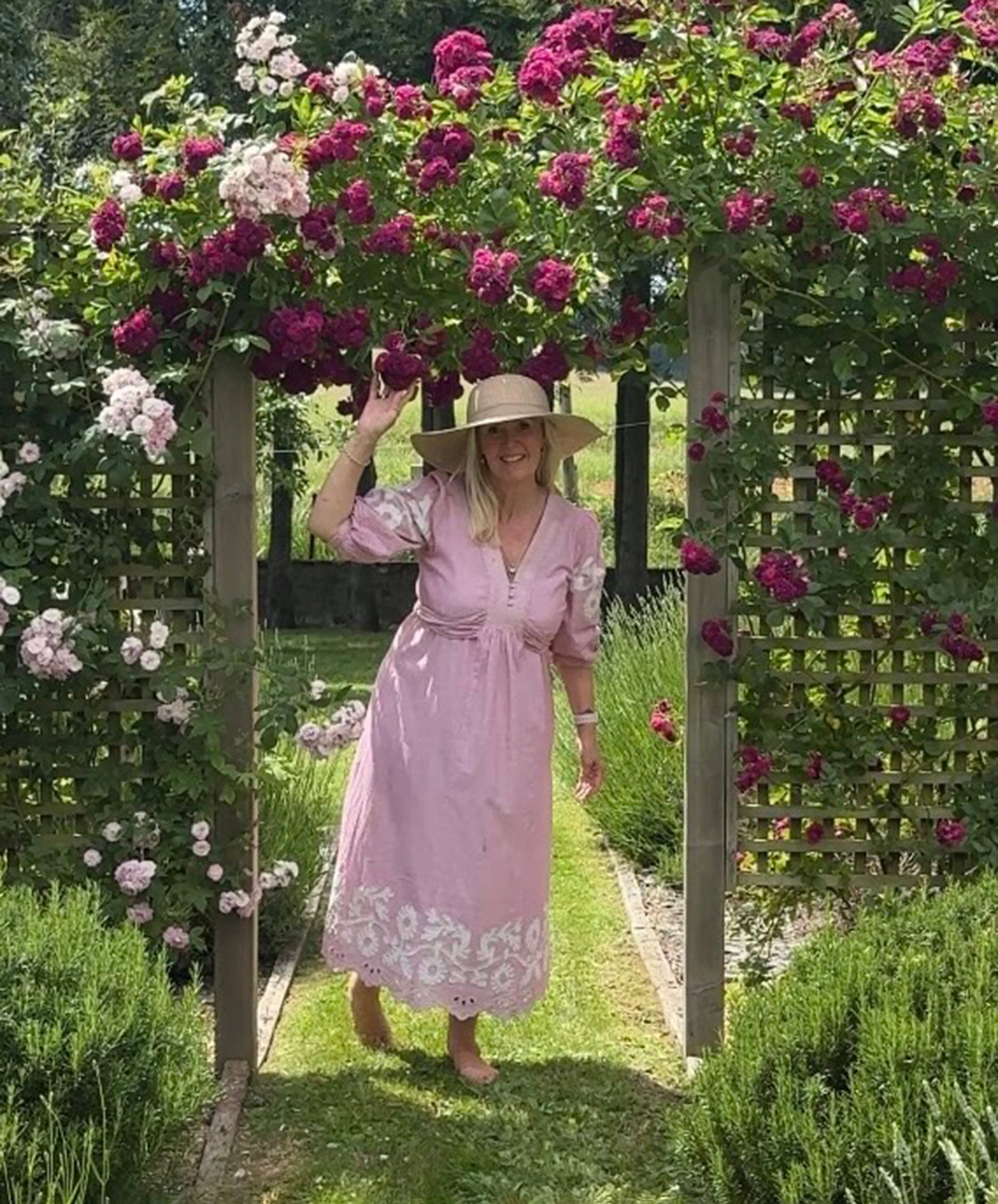 Influencer @shutters_and_stone wearing the Begonia pink dress and hat standing under a floral archway in a garden.