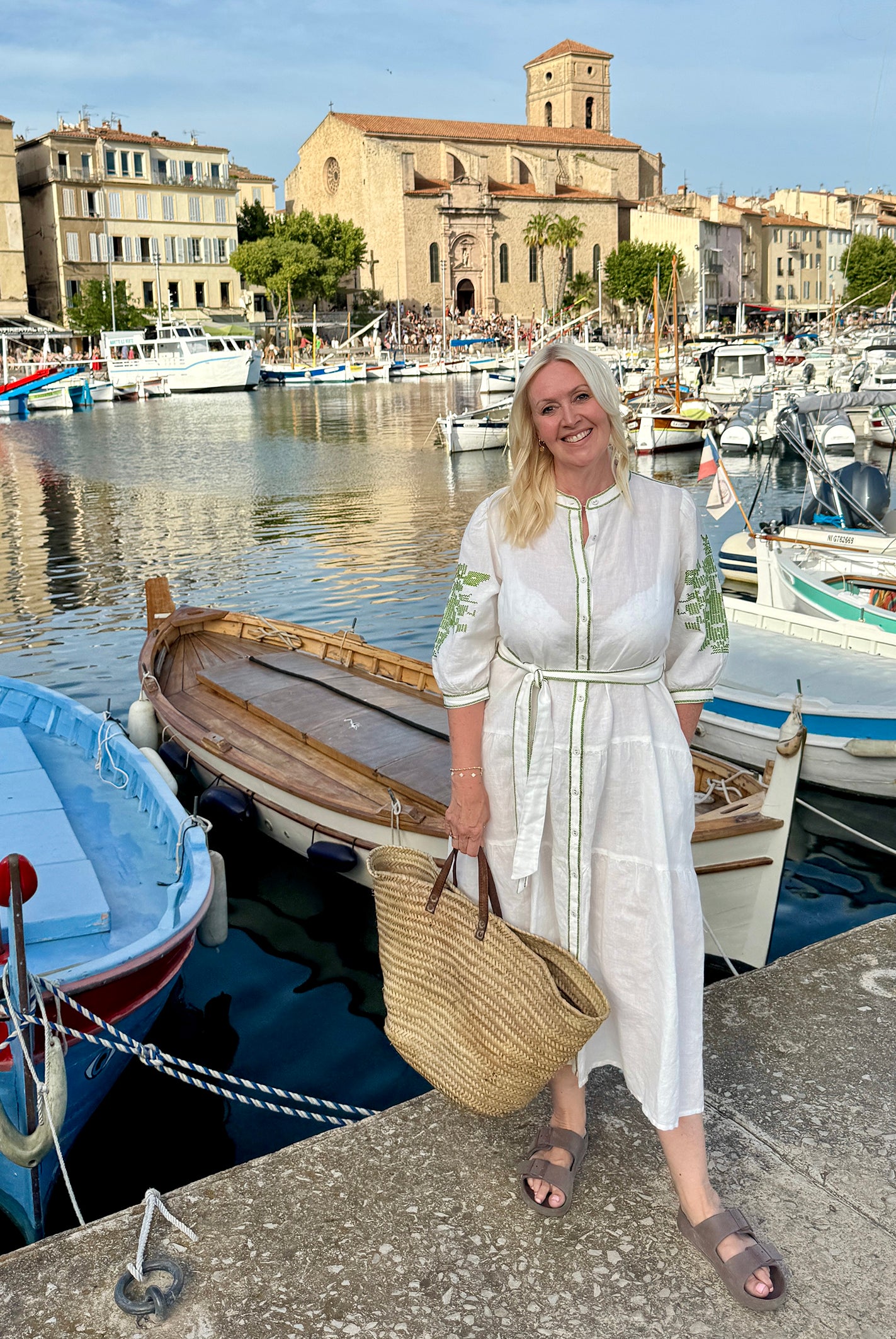 Influencer @shutters_and_stone wearing the Pine white dress with olive green embroidery, stood next to an old marina.