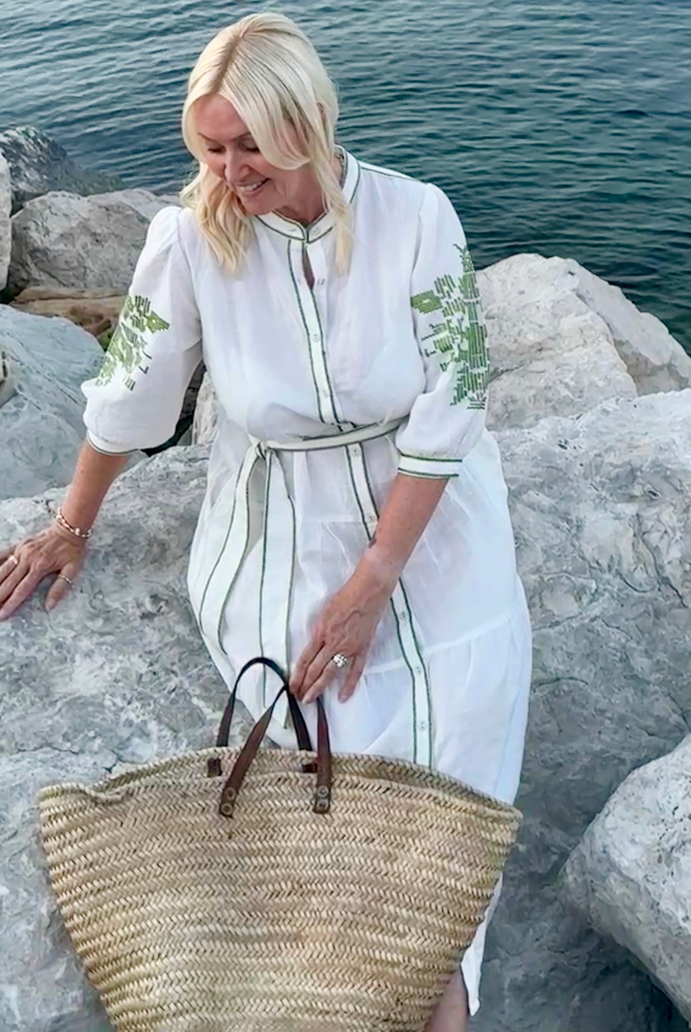 Influencer @shutters_and_stone wearing the Pine white dress with olive green embroidery, sitting on a rock by the sea with a straw bag in hand.