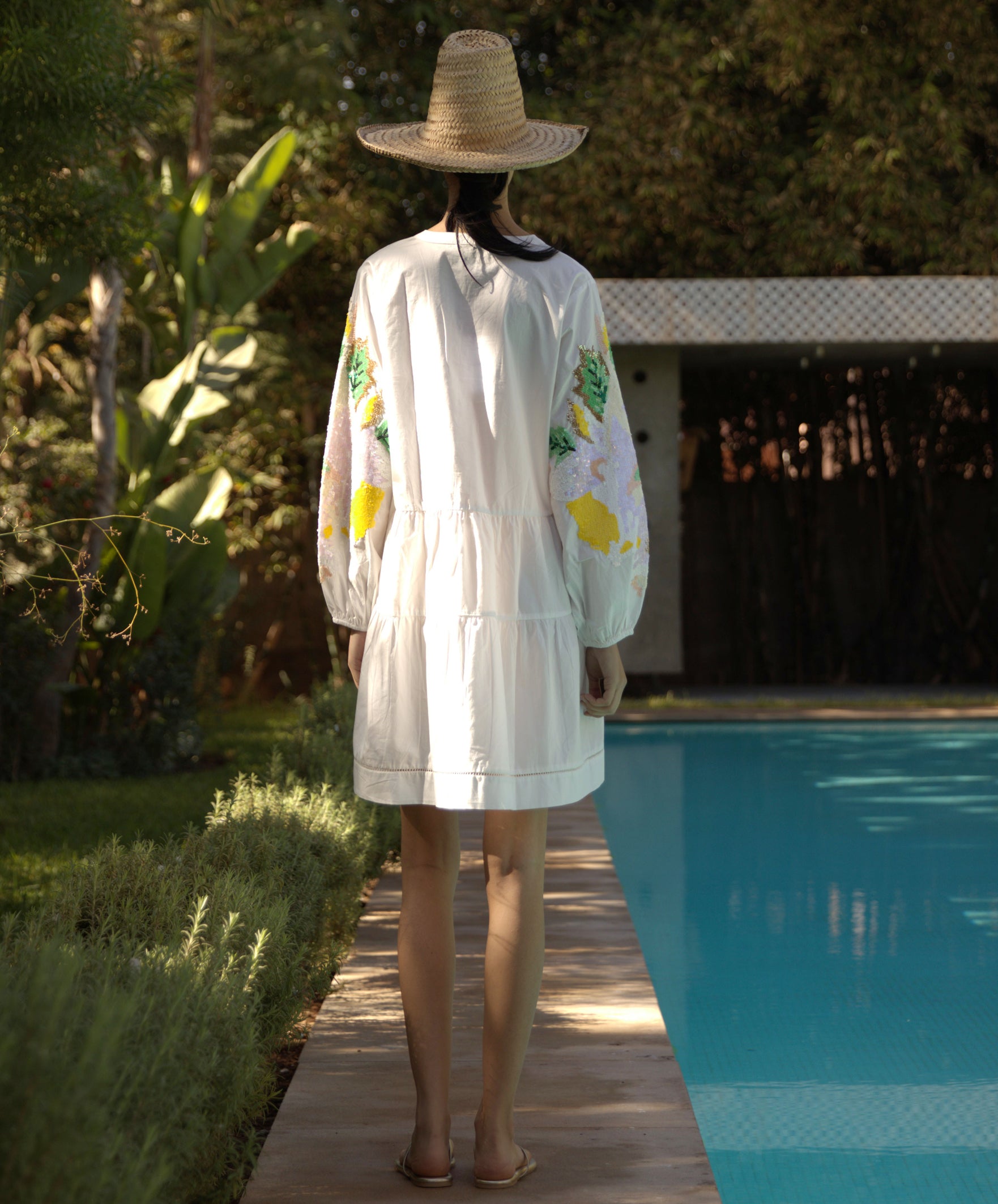 Back view of a woman wearing a Beech white dress with sequin decorated sleeves by a poolside