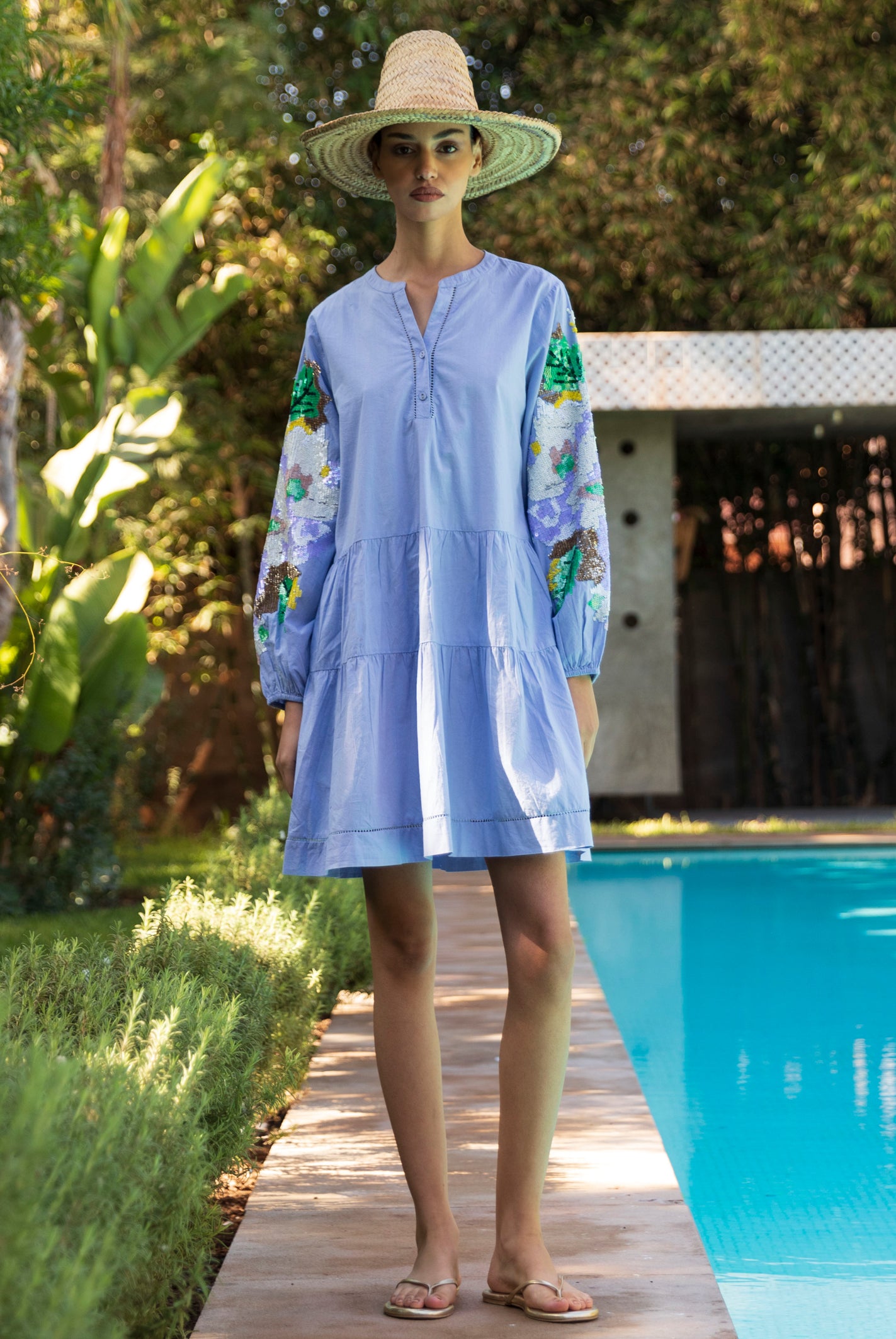 Woman in a light blue Beech dress with sequin decorated sleeves standing by a poolside.