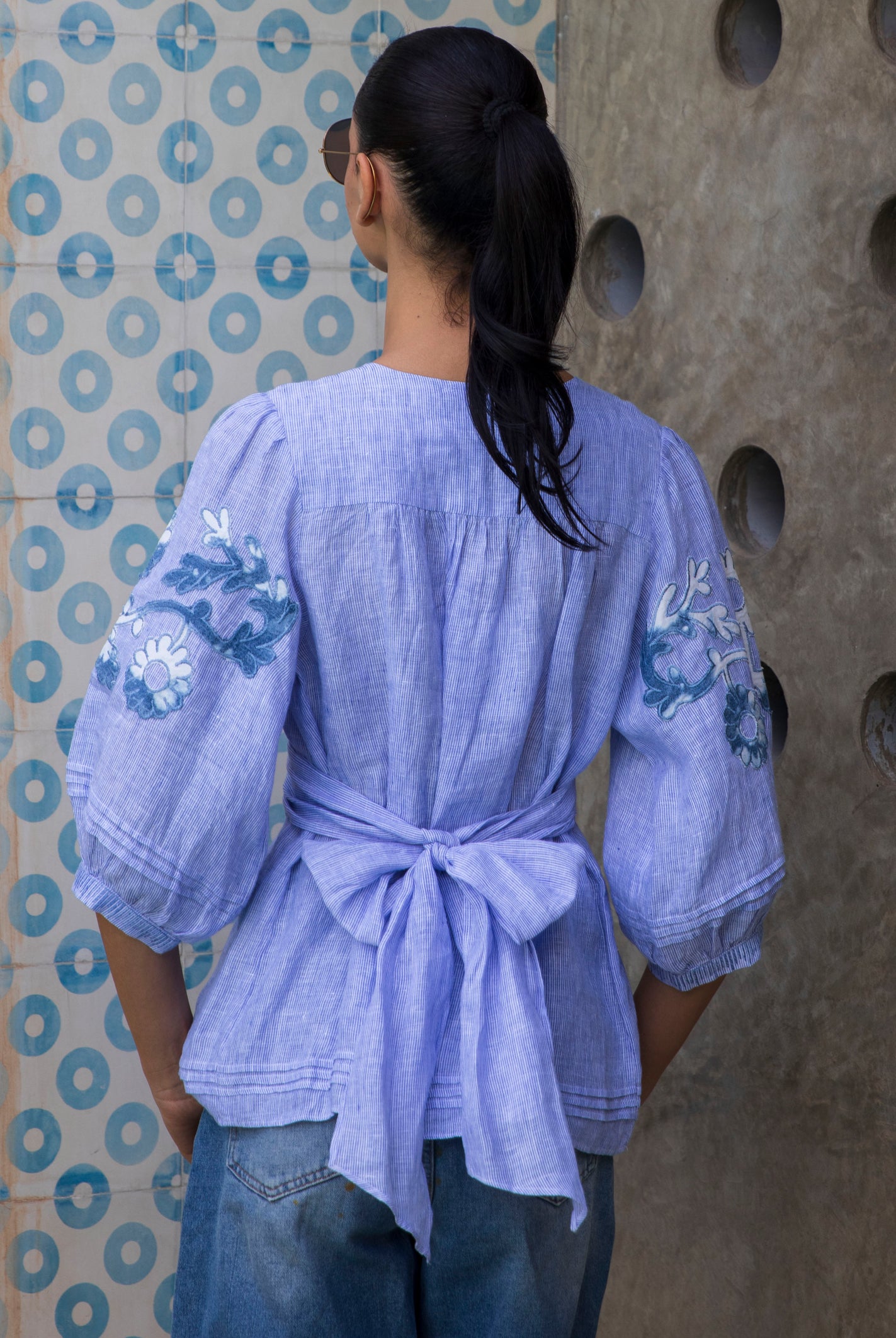 Woman wearing a blue striped Blossom embroidered blouse against a patterned wall.