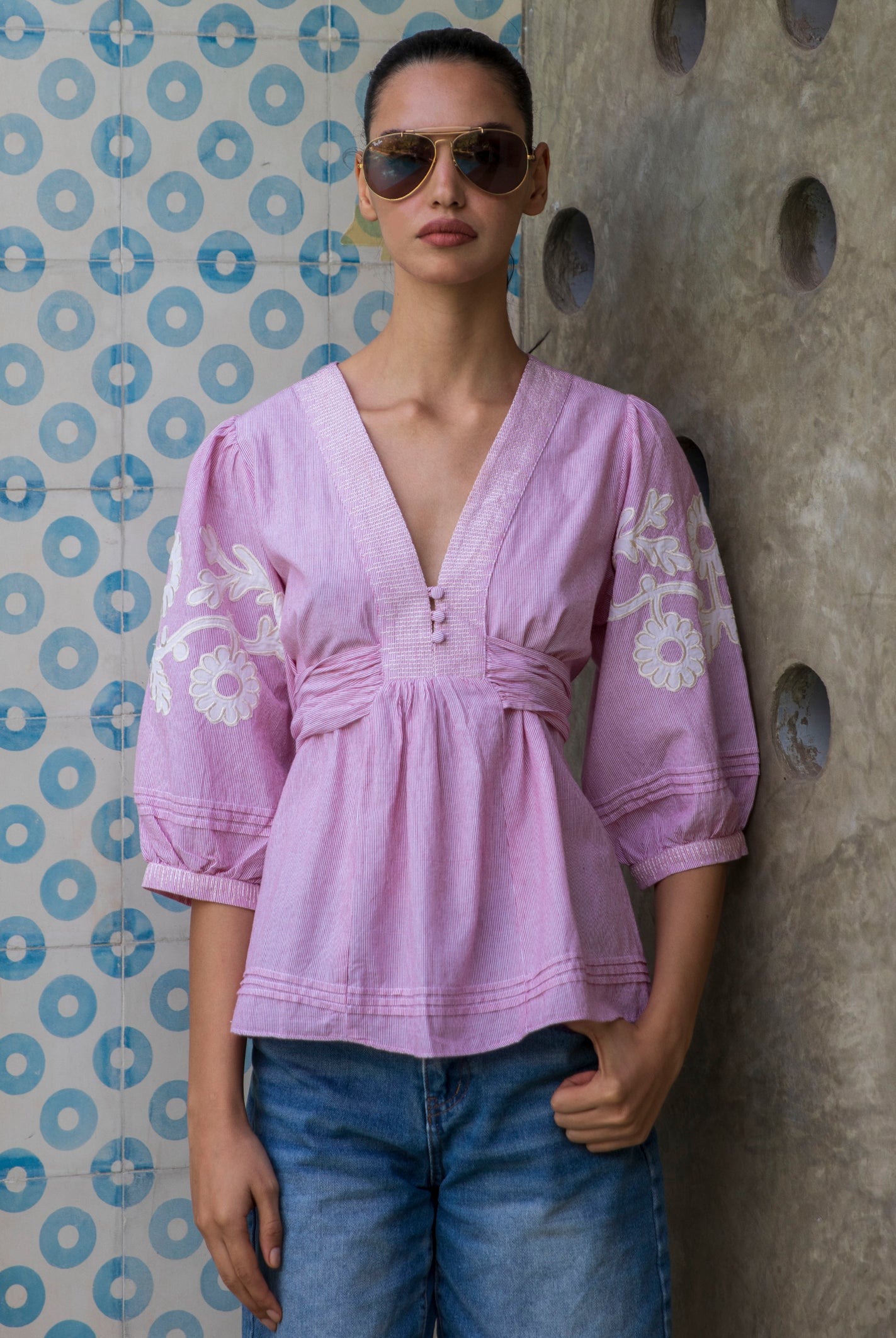 Woman wearing a red striped Blossom blouse with floral patterns and sunglasses, standing against a patterned wall.