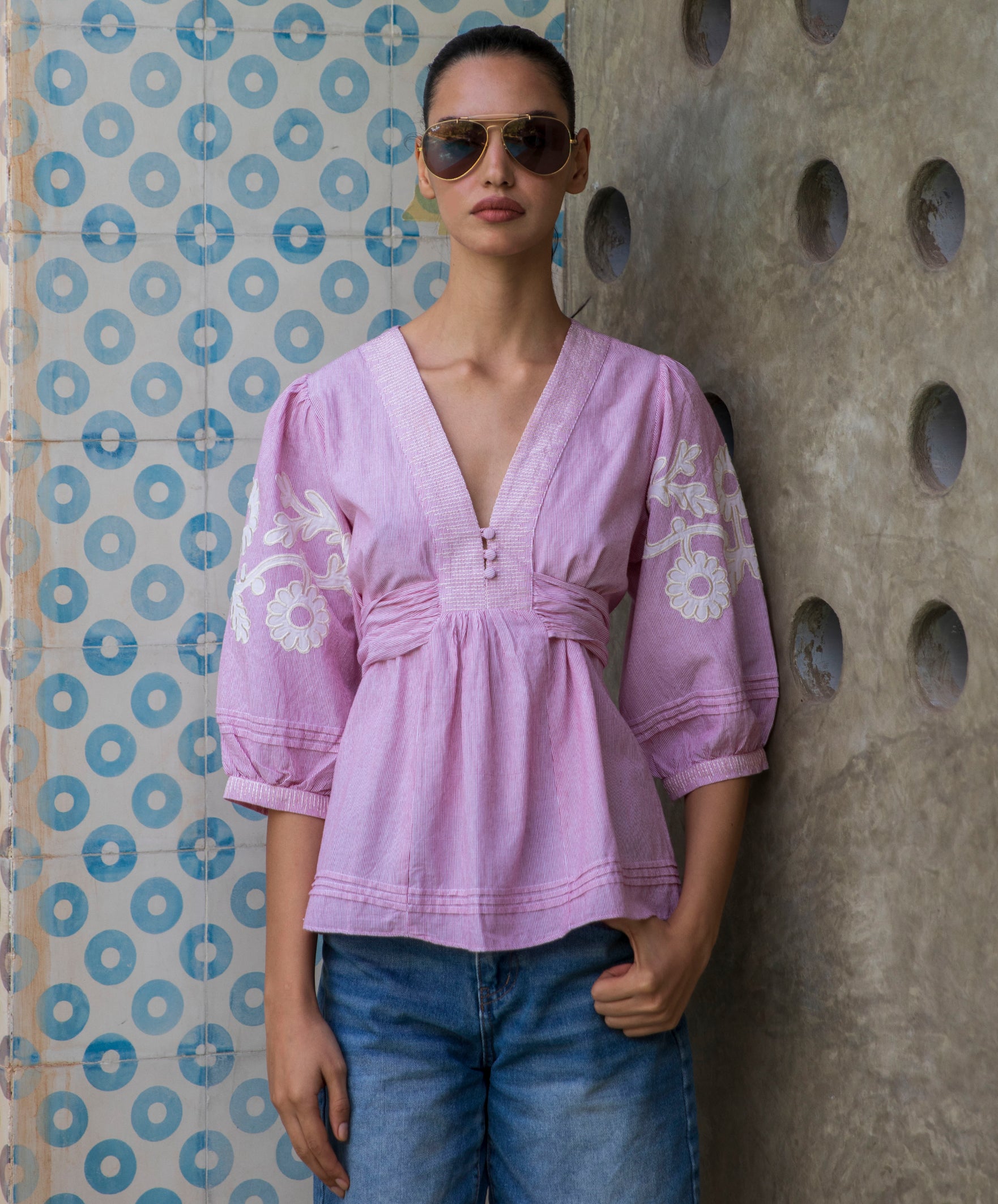 Woman wearing a red striped Blossom blouse with floral patterns and sunglasses, standing against a patterned wall.