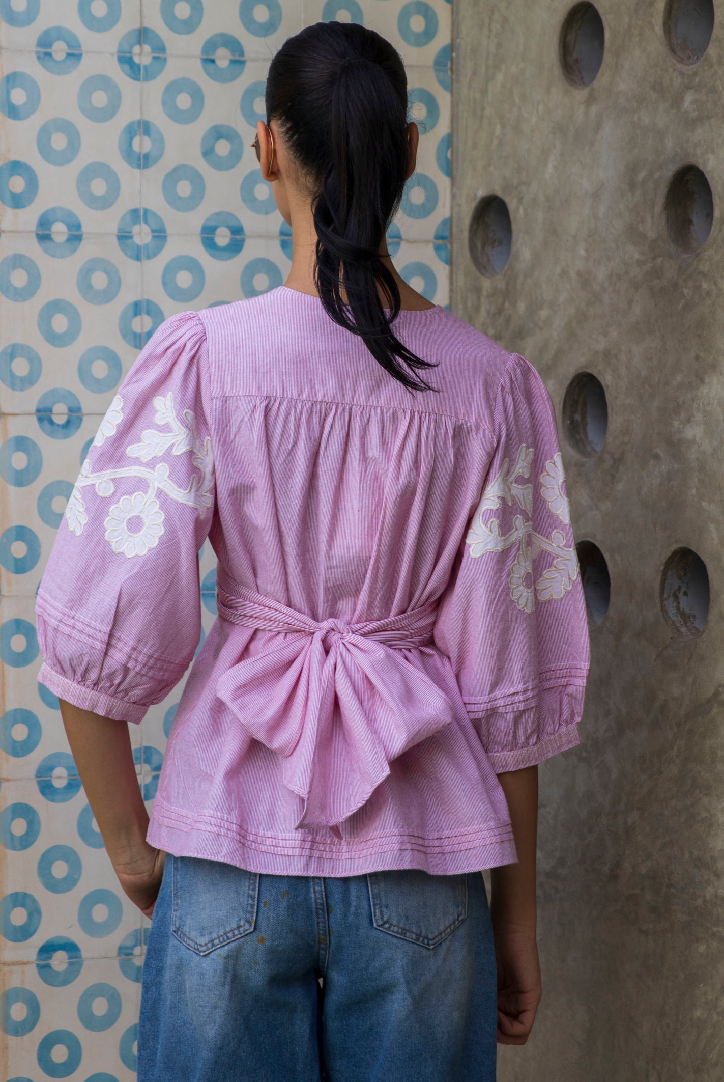 Woman wearing a red striped Blossom blouse with floral patterns and blue jeans against a patterned wall.