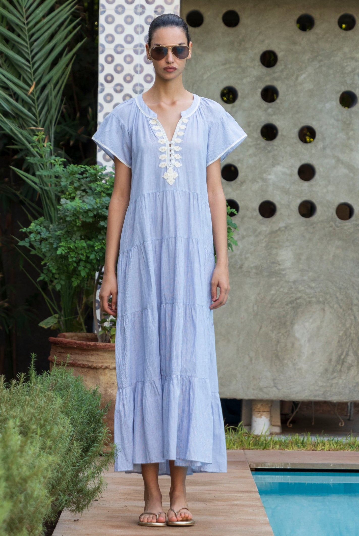 Woman in a striped blue Camelia dress with passementerie embroidery standing by a pool