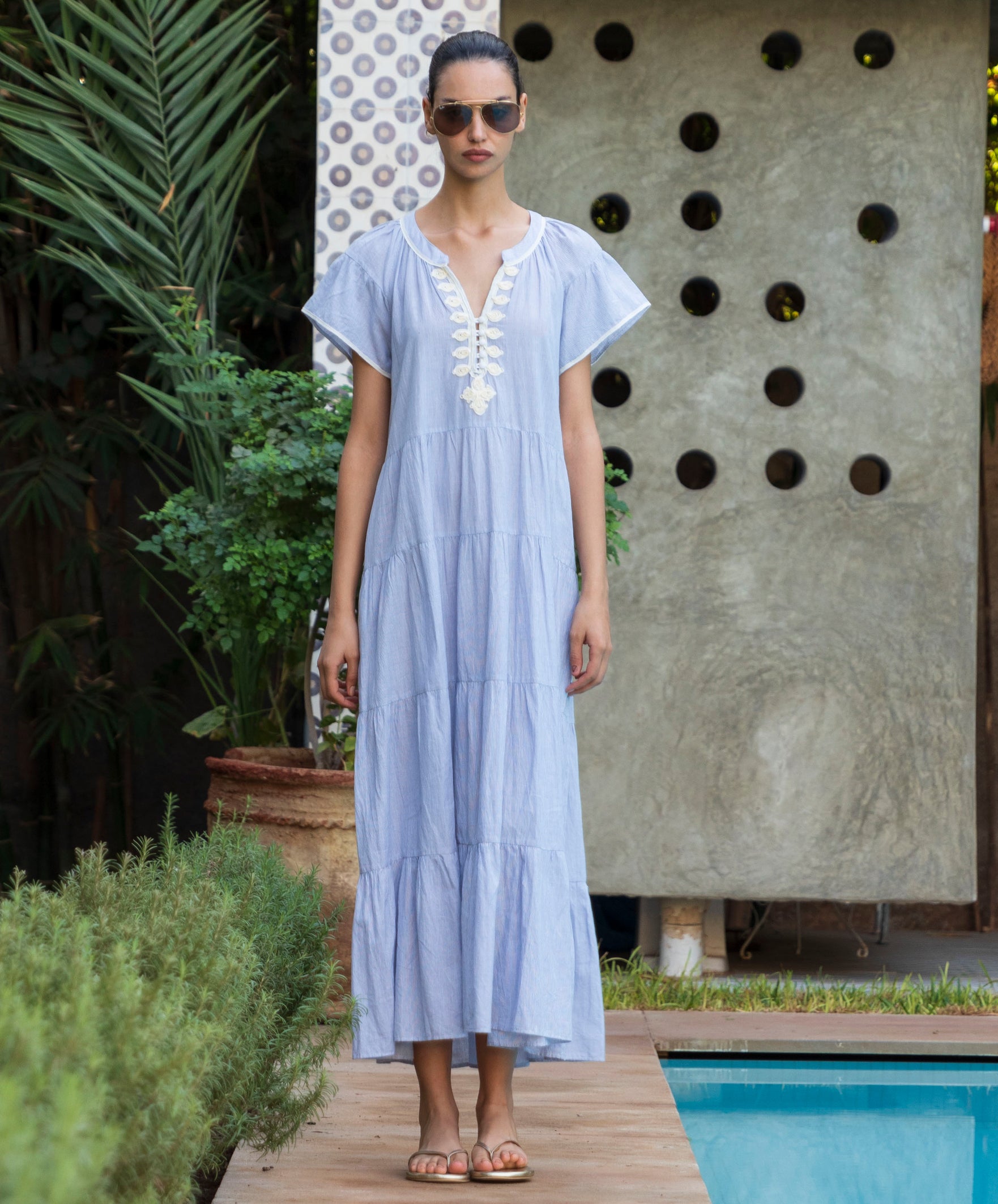 Woman in a striped blue Camelia dress with passementerie embroidery standing by a pool