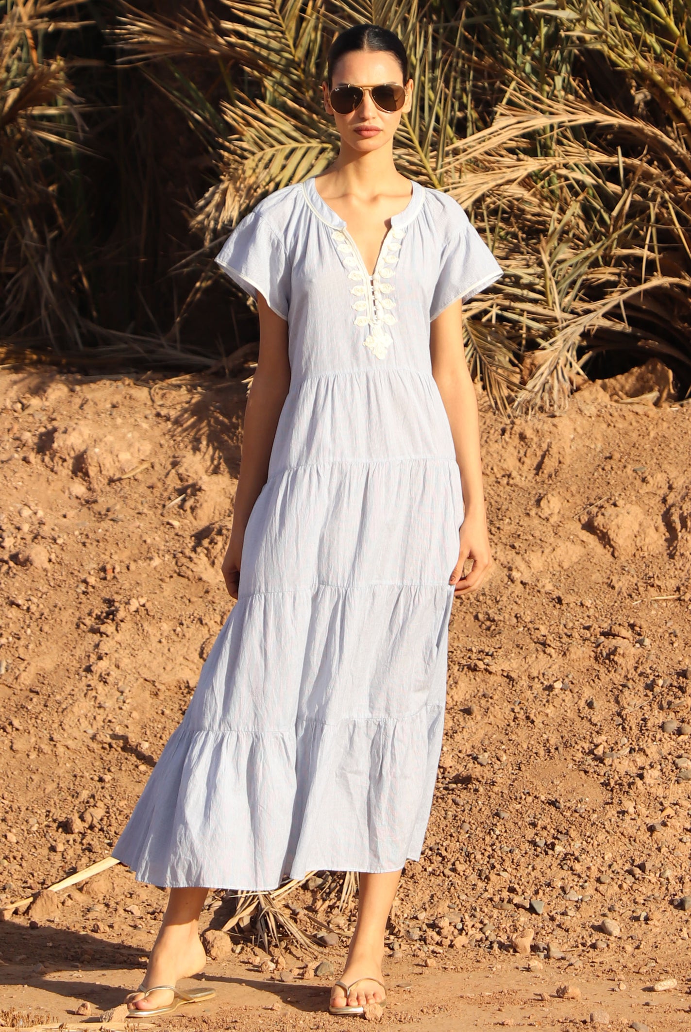 Woman in a striped blue Camelia dress standing in a desert-like setting with palm trees.