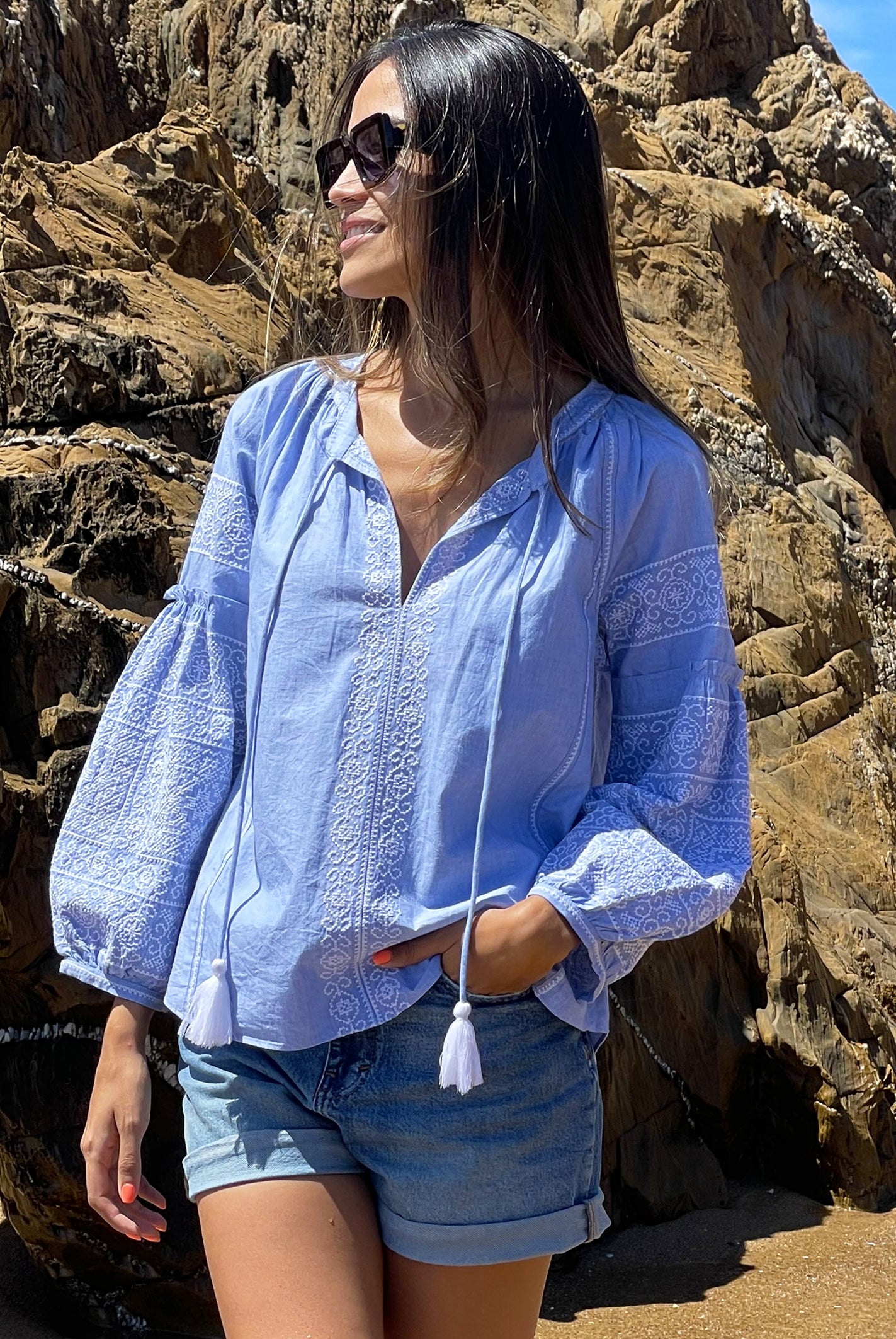 A model stood in front of a rock wearing a Rose and Rose blue cotton Capri top, denim shorts and sunglasses.  