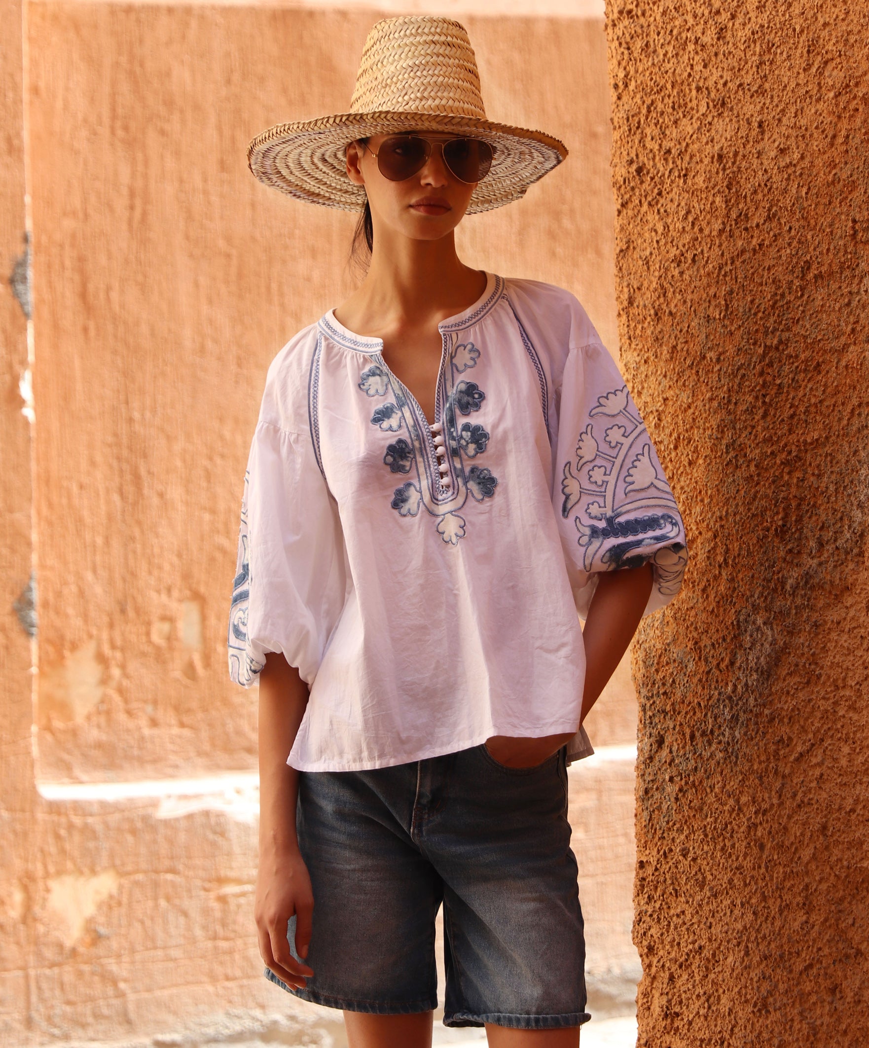 Woman wearing a white blouse with denim applique embroidery and denim shorts with a straw hat against a warm-toned wall.