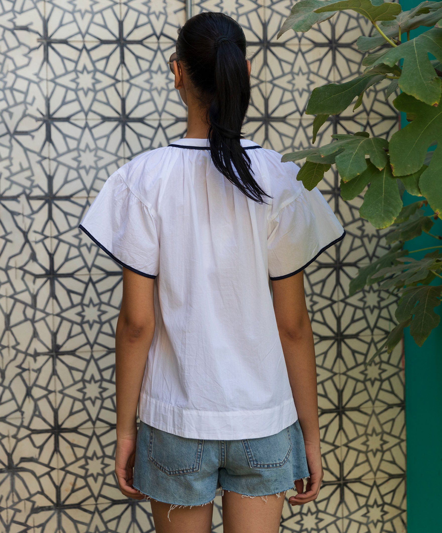Woman wearing a white Clematis blouse with Navy passementerie embroidery and denim shorts standing against a patterned wall.