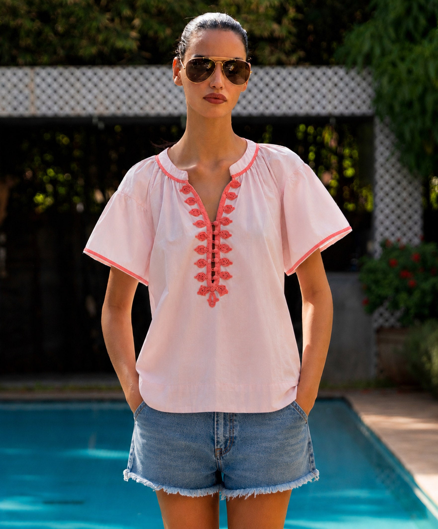 Woman wearing a light pink blouse with coral embroidery and denim shorts by a pool.