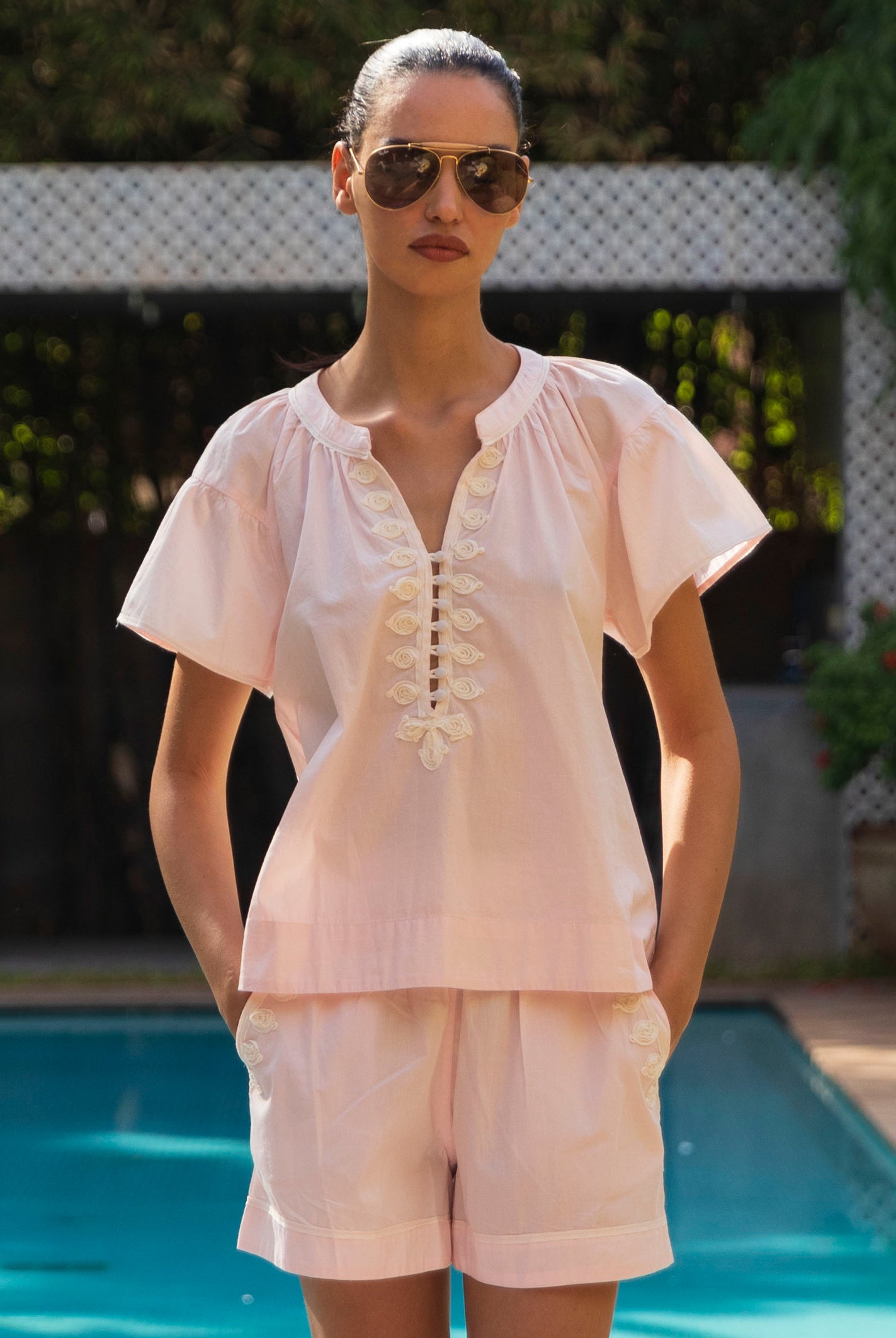 Woman wearing a pale pink Clematis blouse with matching shorts and sunglasses by a pool