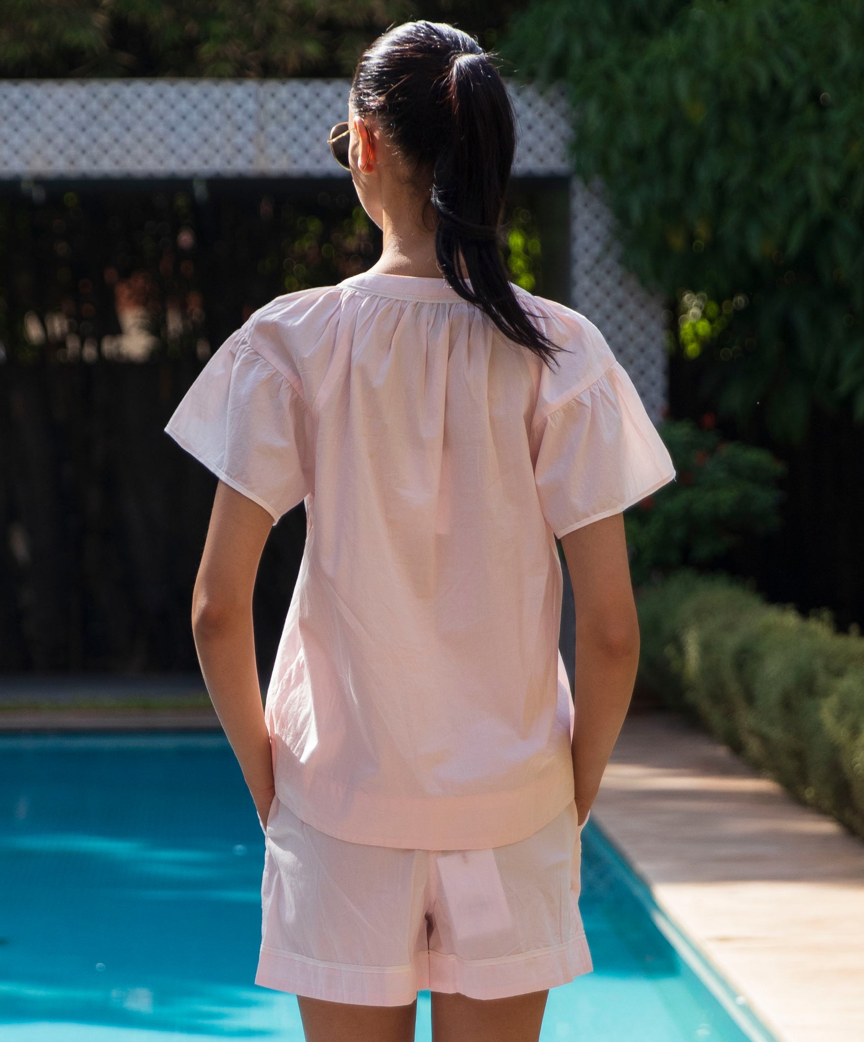 Back view of a women wearing a light pink Clematis top and shorts by a poolside