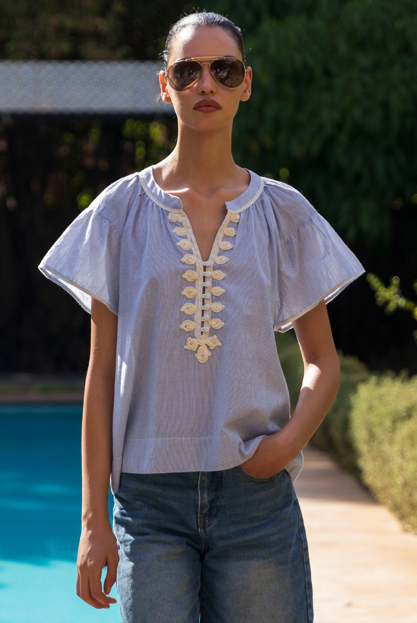 Woman wearing a blue striped blouse with passementerie embroidery details and sunglasses by a poolside.