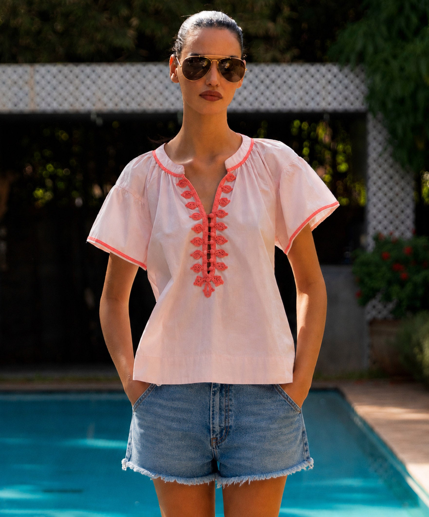 Woman wearing a light pink blouse with coral embroidery and denim shorts by a pool.
