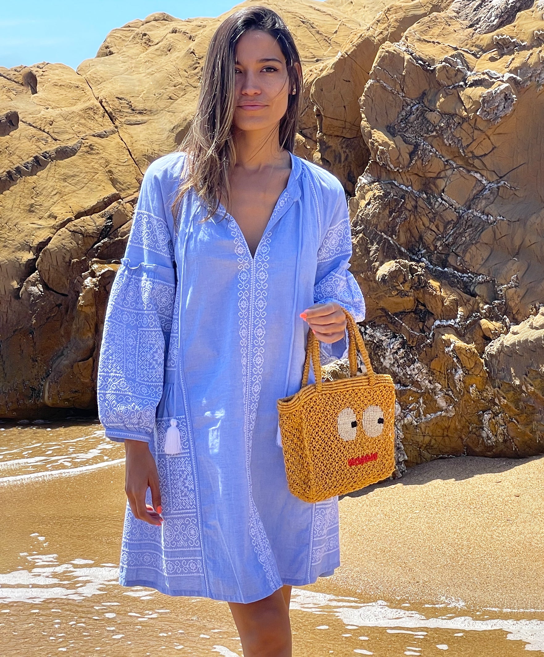 A model stood on a beach wearing a Rose and Rose blue Como dress and holding a Jacksons jute bag. 