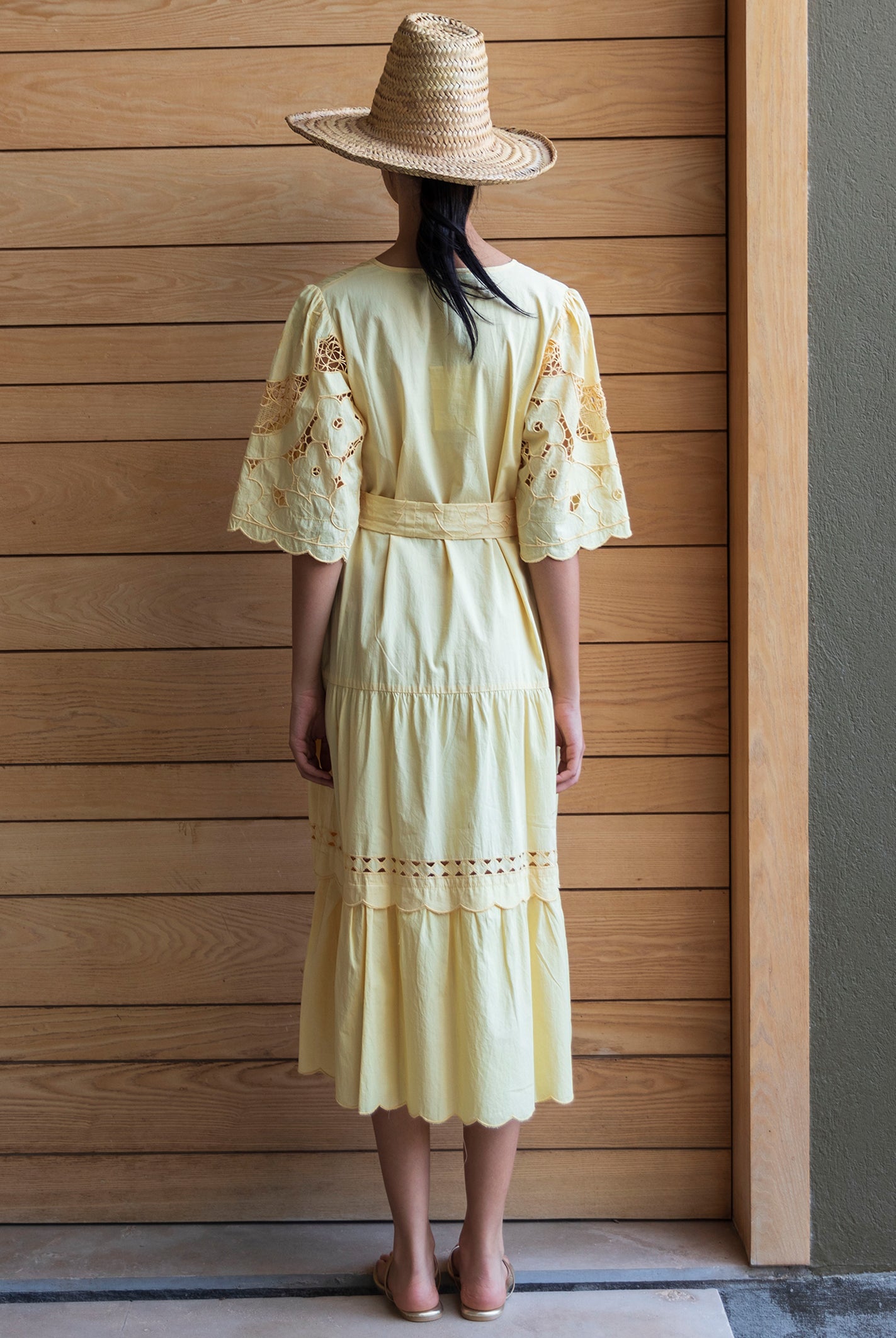 Woman wearing a light yellow embroidered dress and a straw hat, standing against a wooden wall.