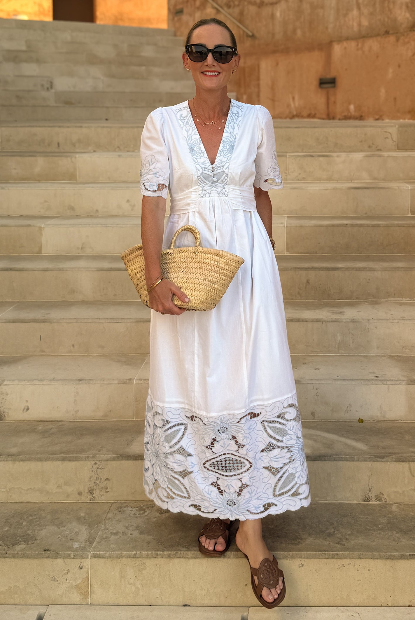 Influencer @asliceofsuffolk stands on outdoor stone steps wearing a white maxi dress with blue embroidery, holding a woven straw tote bag.