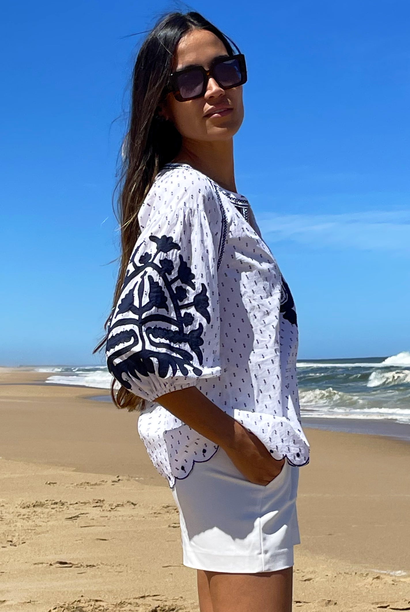 A model stood on a beach wearing a Rose and Rose cotton dobby Ferrara top, white shorts and sunglasses.