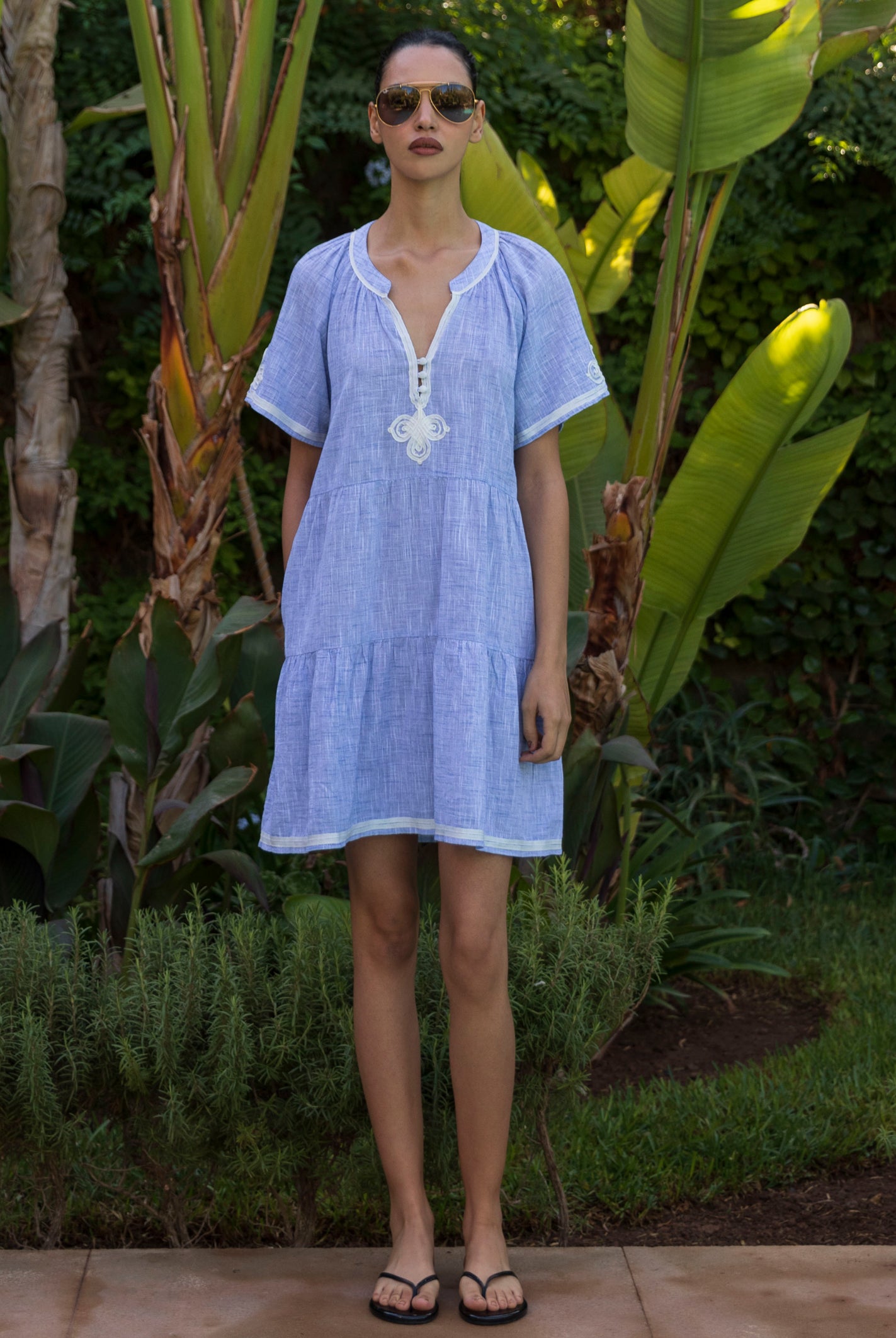 Woman wearing a light blue Flora dress with white passementerie decoration standing in a tropical garden setting