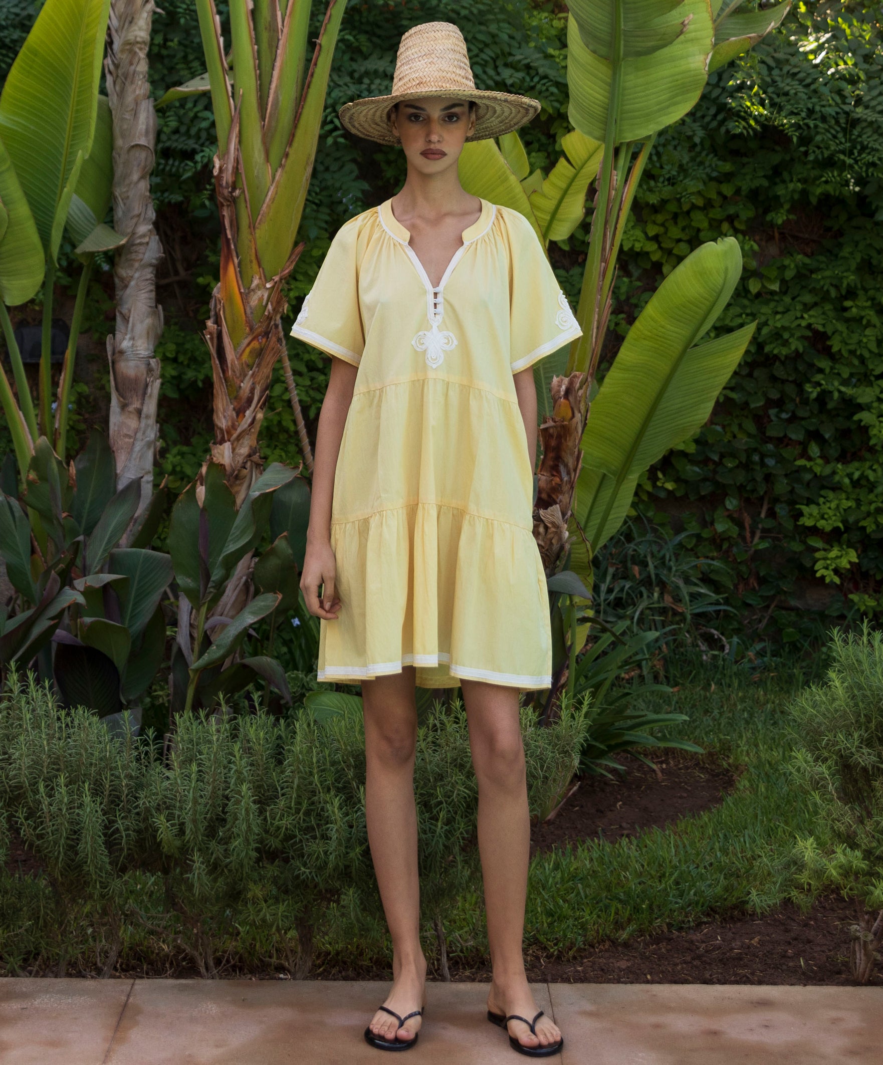 Woman wearing a pale yellow Flora dress and straw hat standing in a tropical garden setting