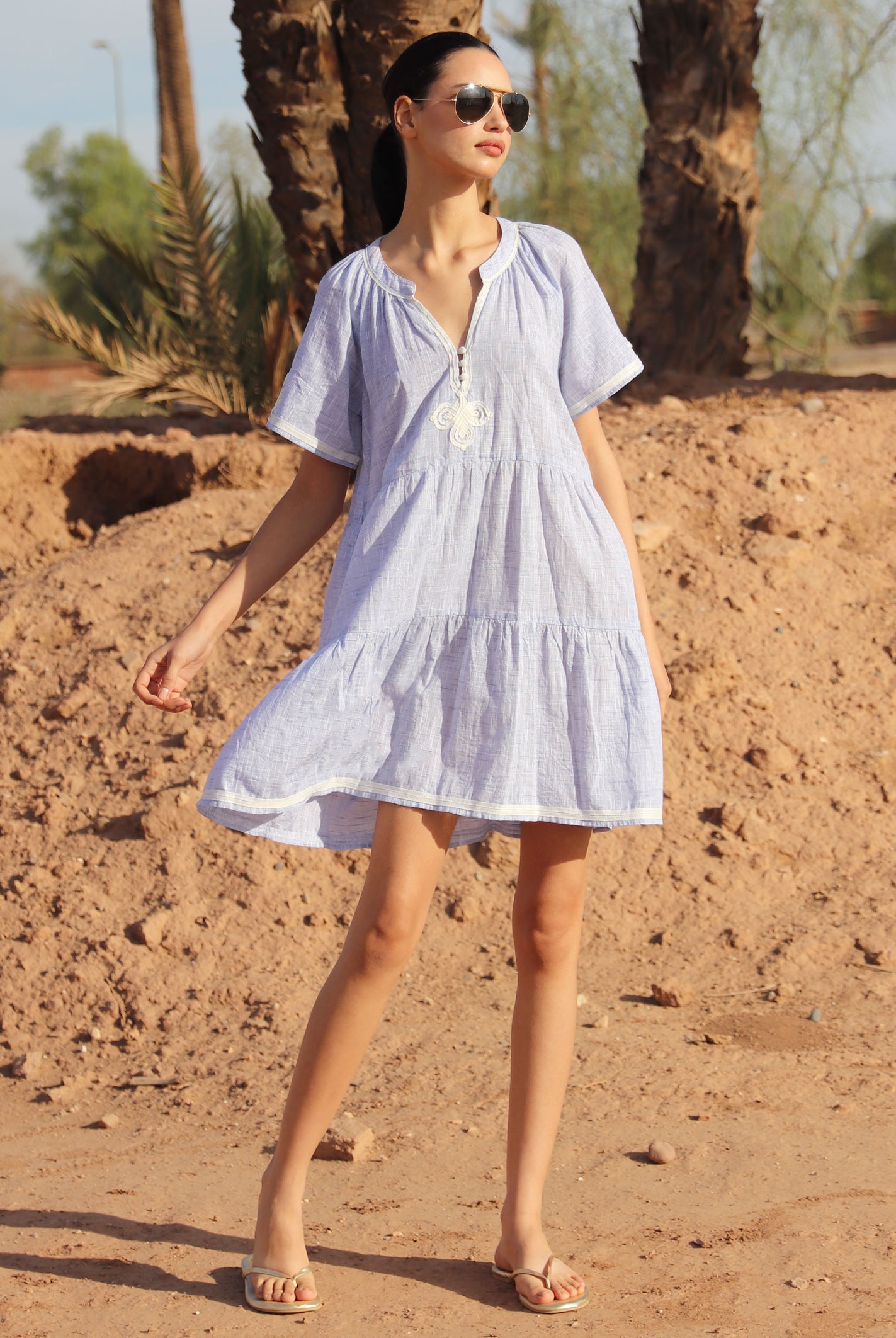 Woman in a light blue Flora dress standing in a desert-like setting with palm trees.
