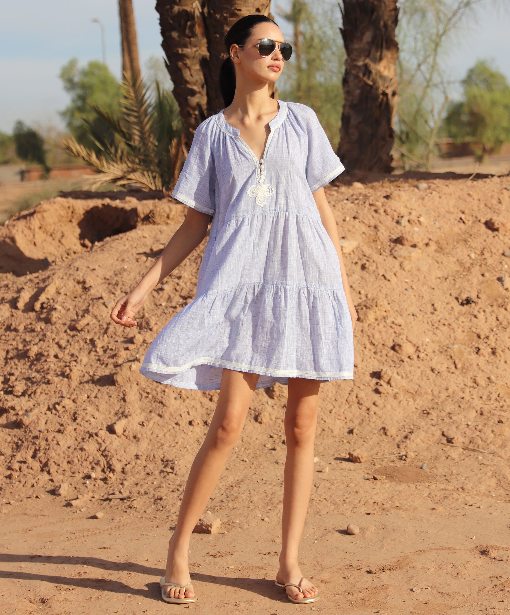Woman in a light blue Flora dress standing in a desert-like setting with palm trees.