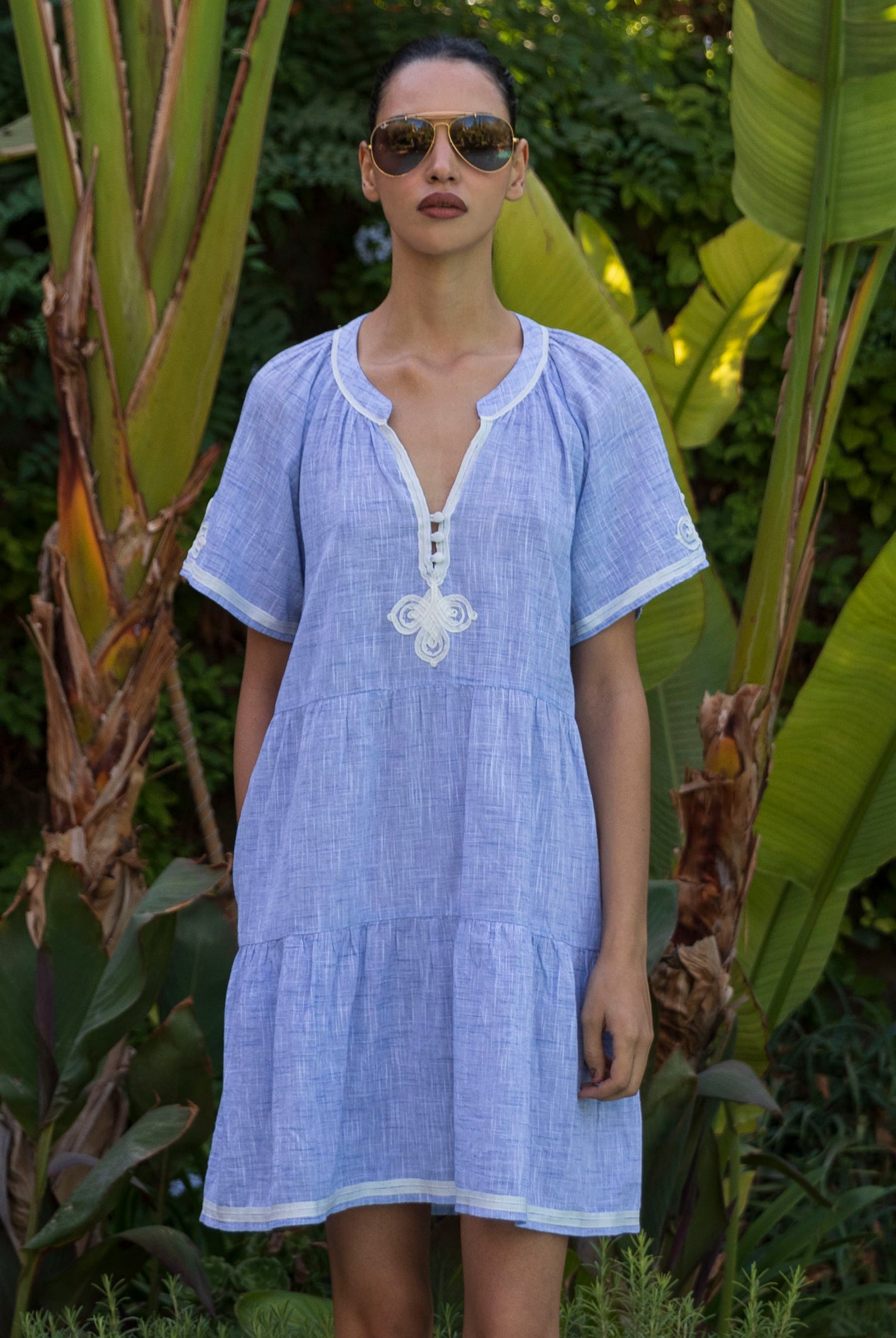 Woman wearing a light blue Flora dress with white passementerie trim standing among tropical plants