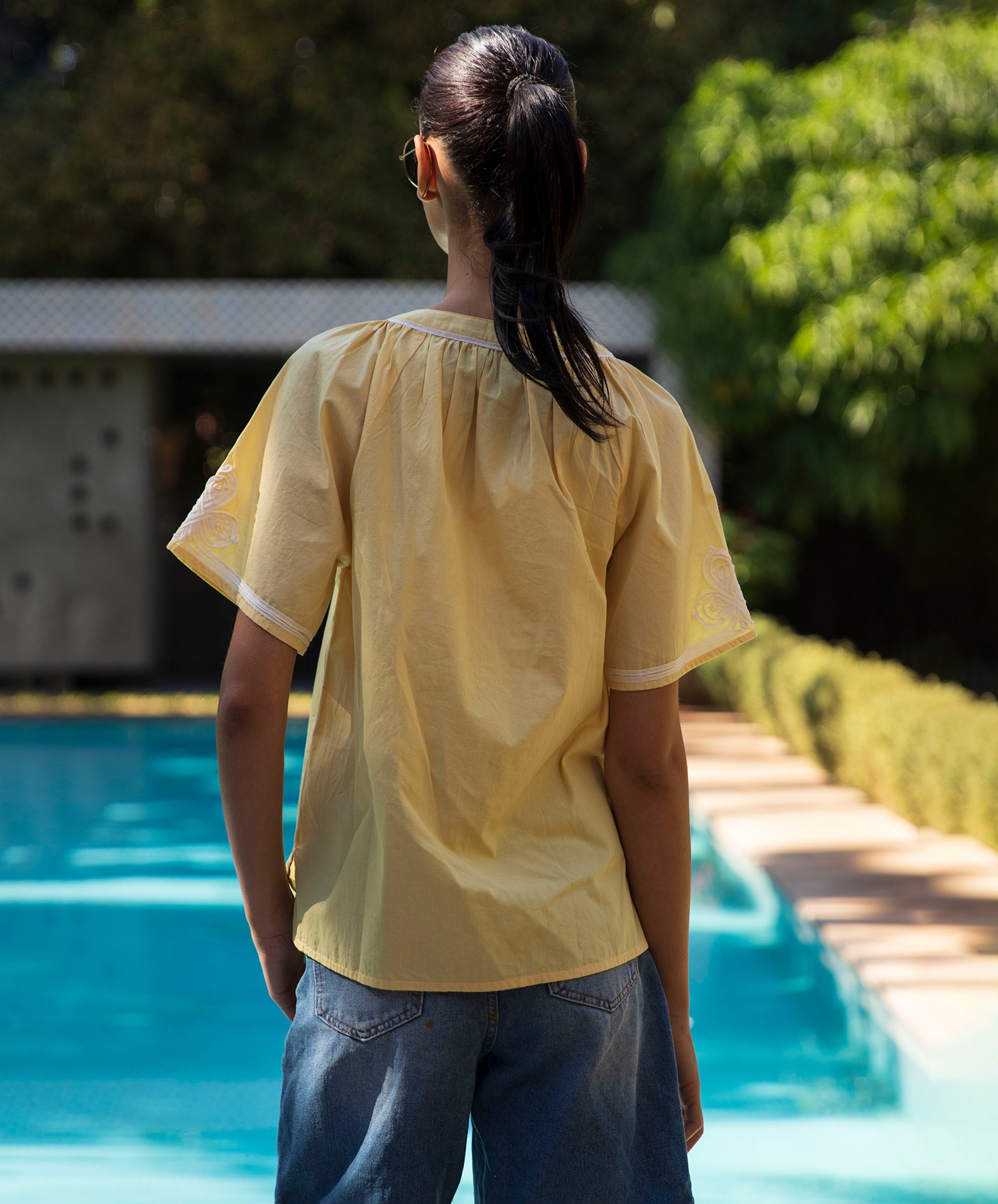 Back view of a woman wearing a pale yellow Foxglove blouse with passementerie by a pool
