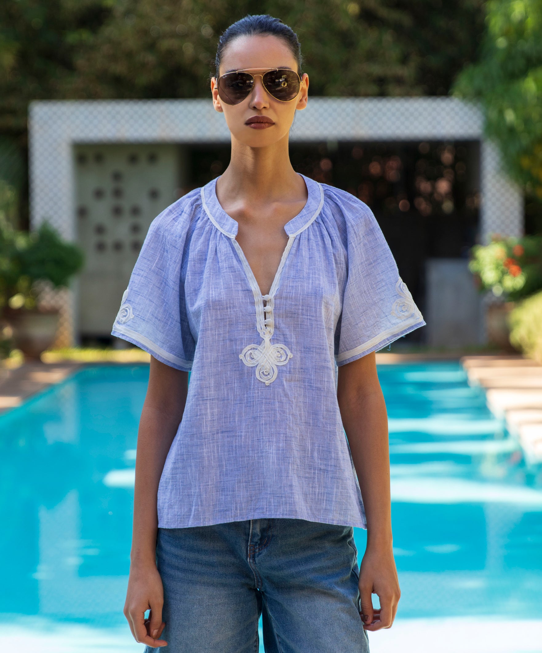 Woman wearing a light blue embroidered Foxglove blouse and sunglasses by a pool