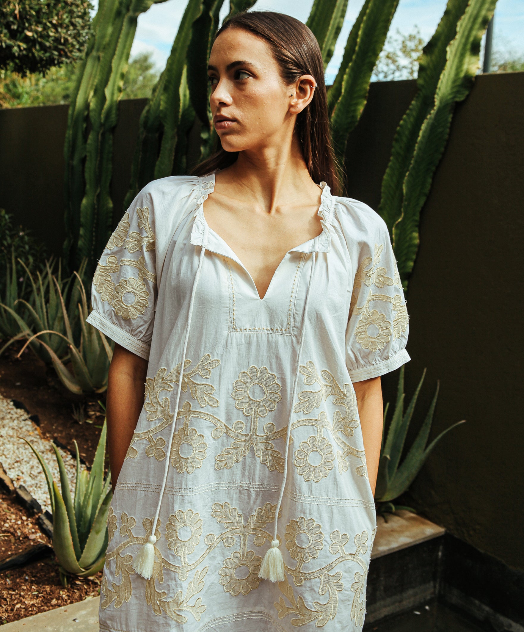 A model wearing a cream Rose and Rose Gloucester cotton dress with gold lurex appliqué decoration stood with her hands in the pockets.