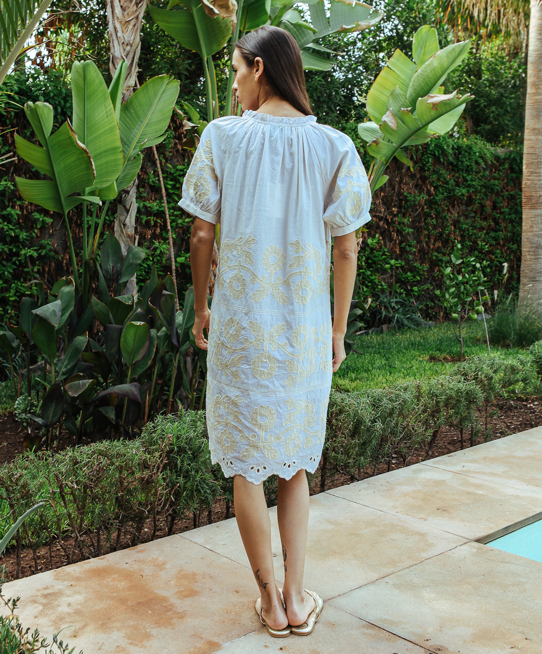 A back view of a model wearing a Rose and Rose Gloucester cream cotton dress with gold lurex appliqué decoration.