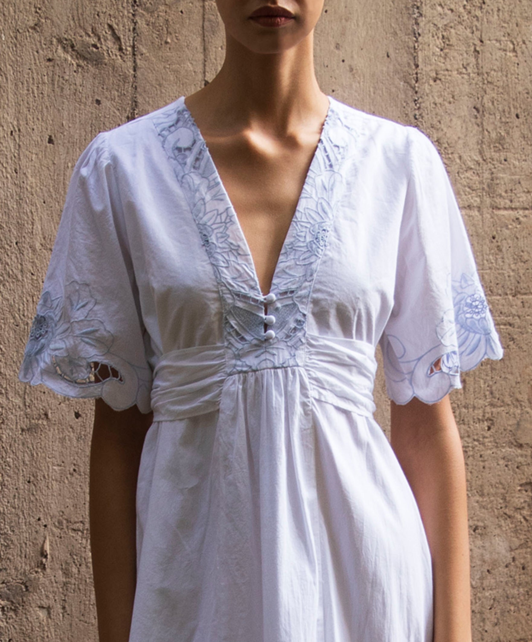 White dress with light blue embroidery details worn by a woman against a beige wall.