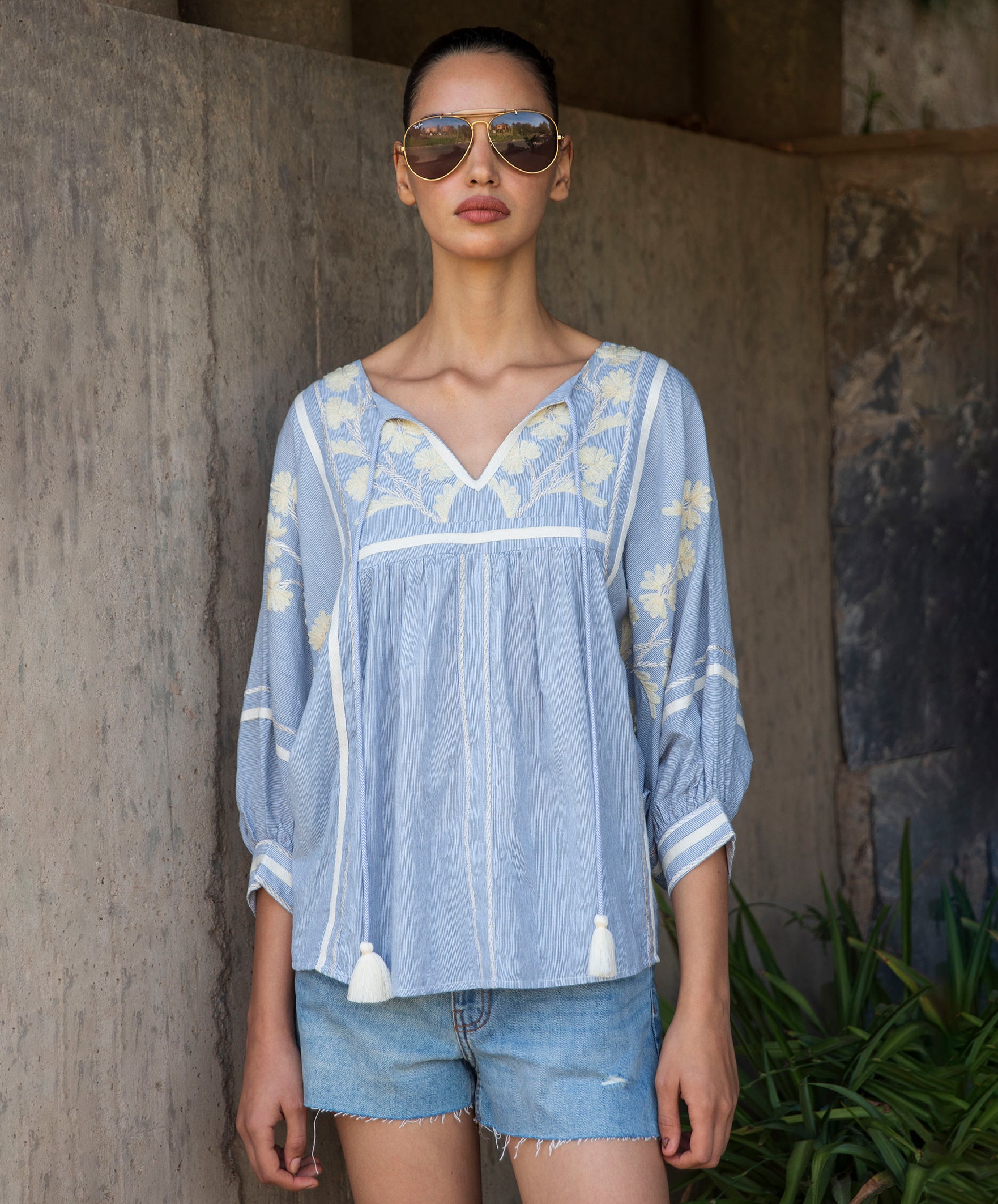 Woman wearing a blue embroidered top and denim shorts with sunglasses, standing against a stone wall.
